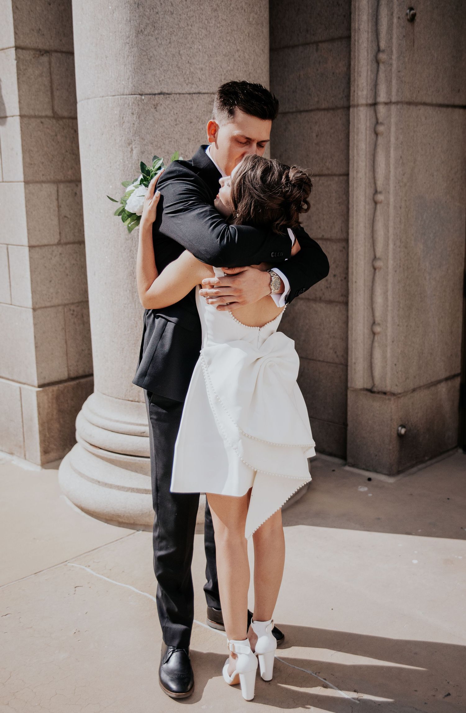A romantic wedding moment captured between a couple embracing in formal attire against stone columns.