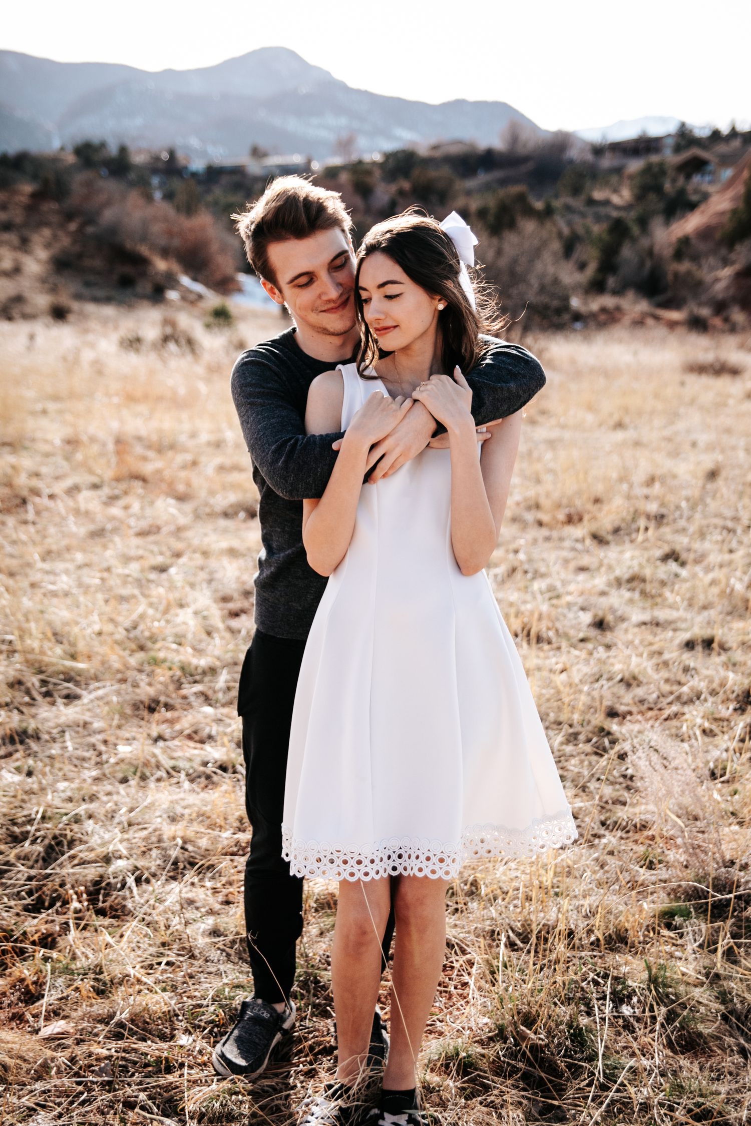 Couple embracing in white dress and dark sweater against mountain backdrop.