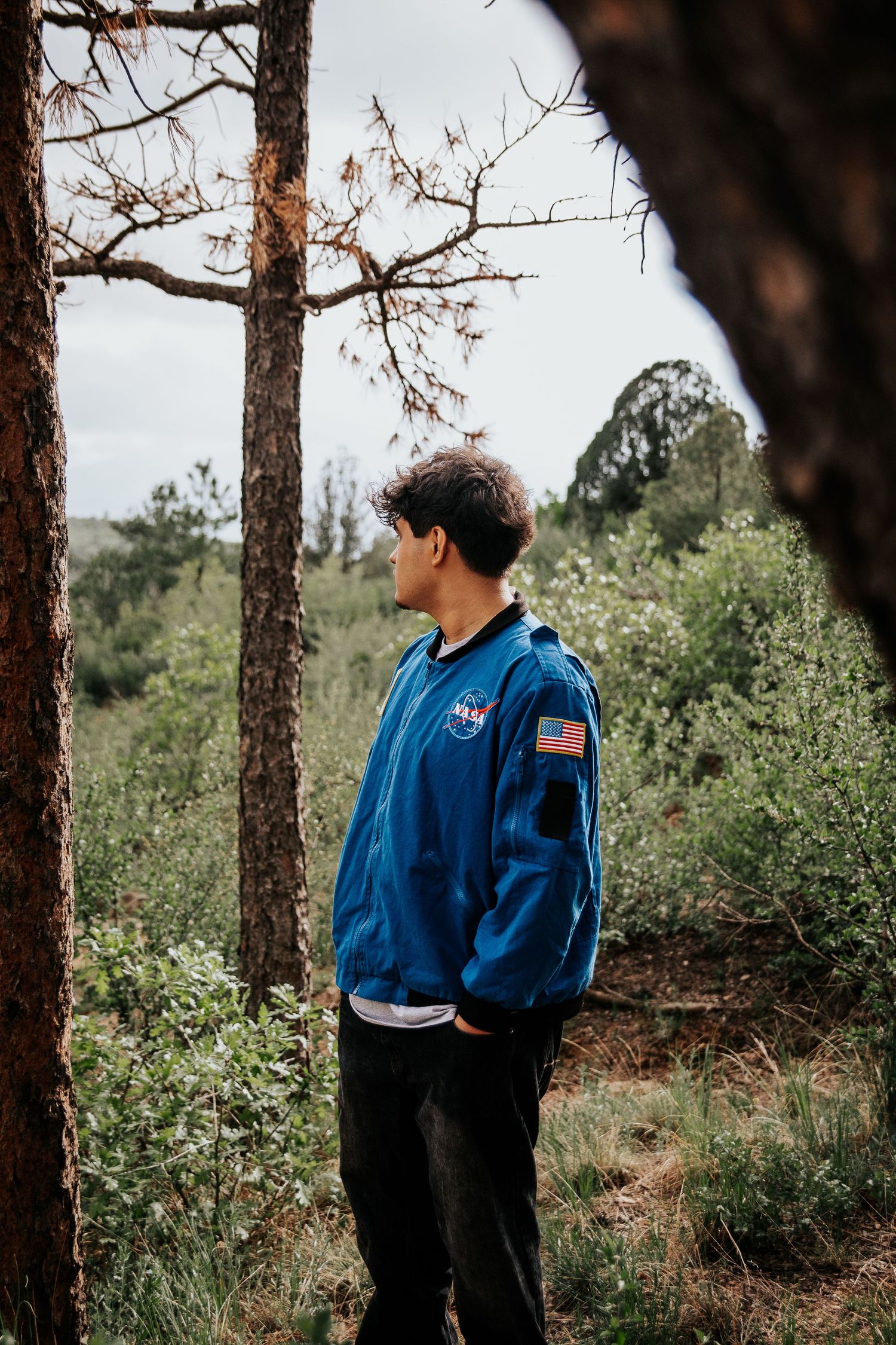 High school senior in a blue NASA jacket stands among pine trees in a natural outdoor setting at Palmer Park in Colorado Springs