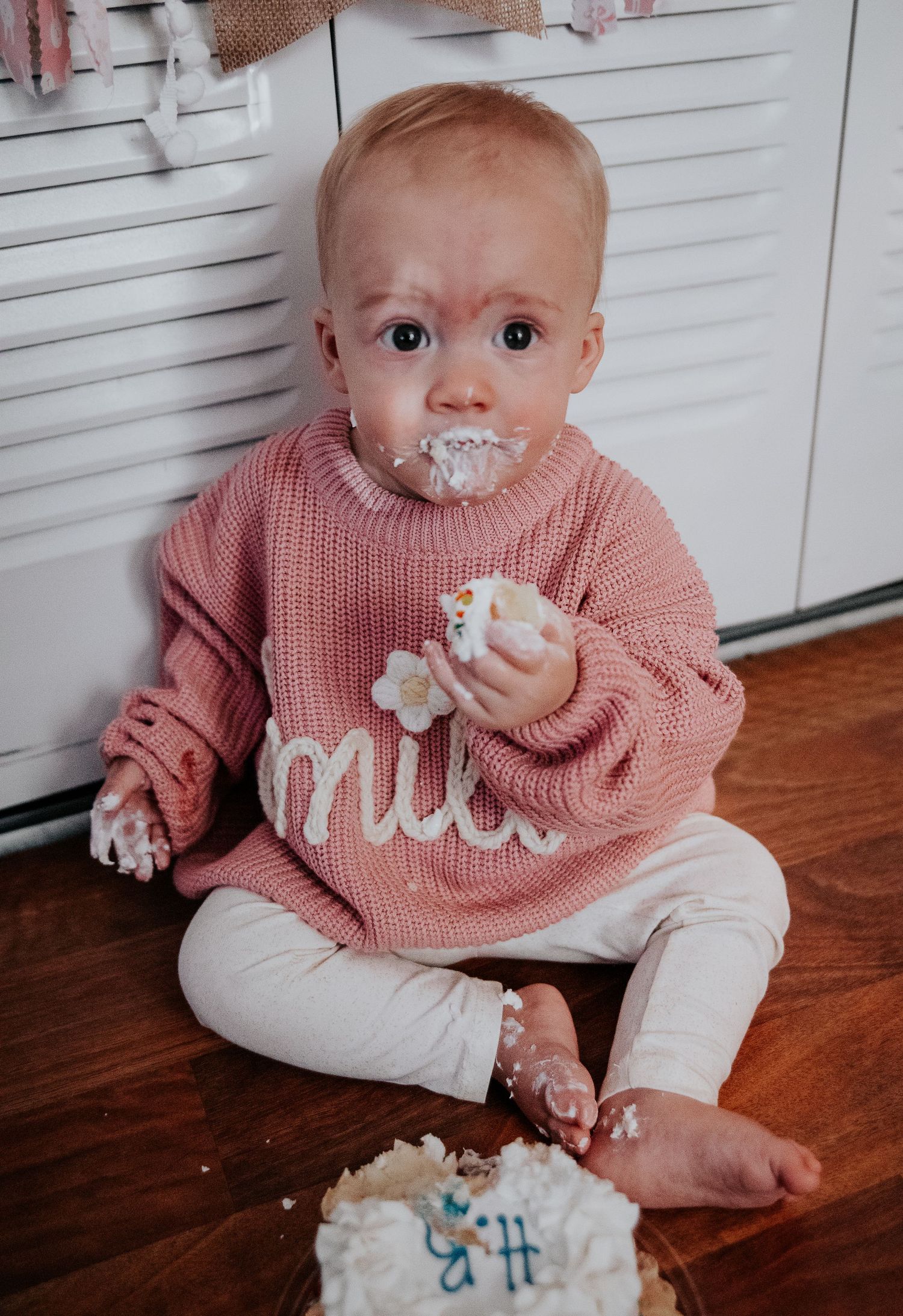 A small child in a pink sweater sits on hardwood floor with cake frosting on their face.