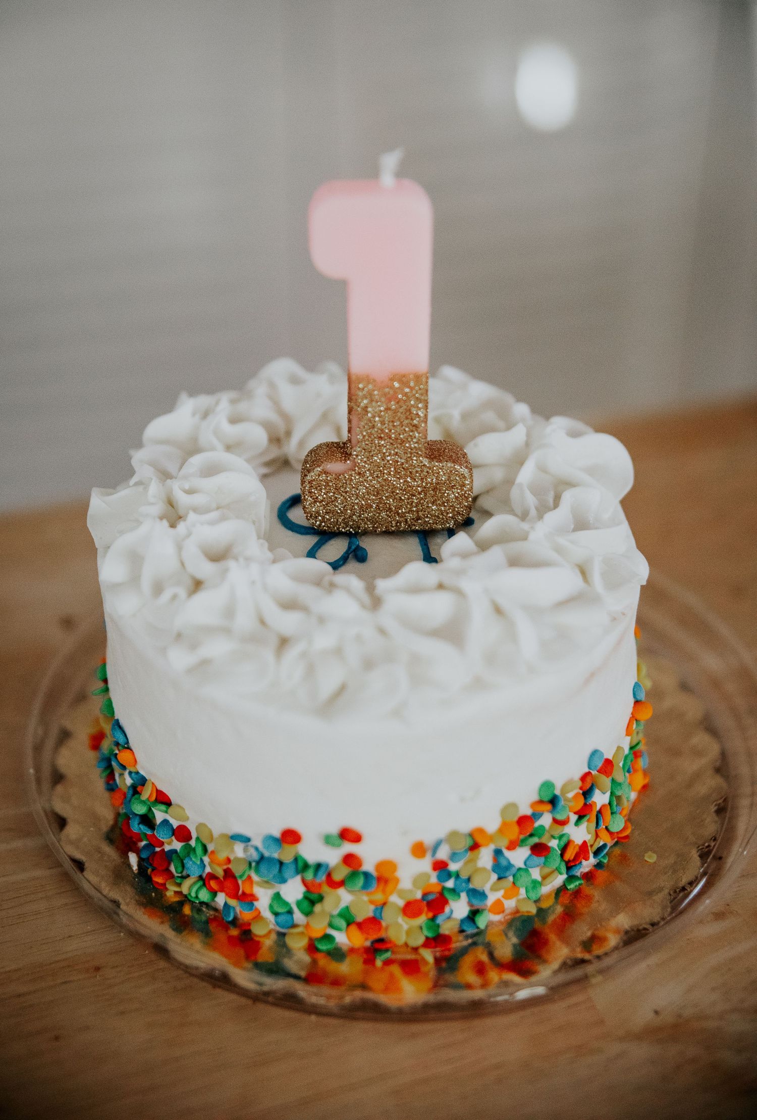 White frosted birthday cake with colorful sprinkles and gold number one candle.