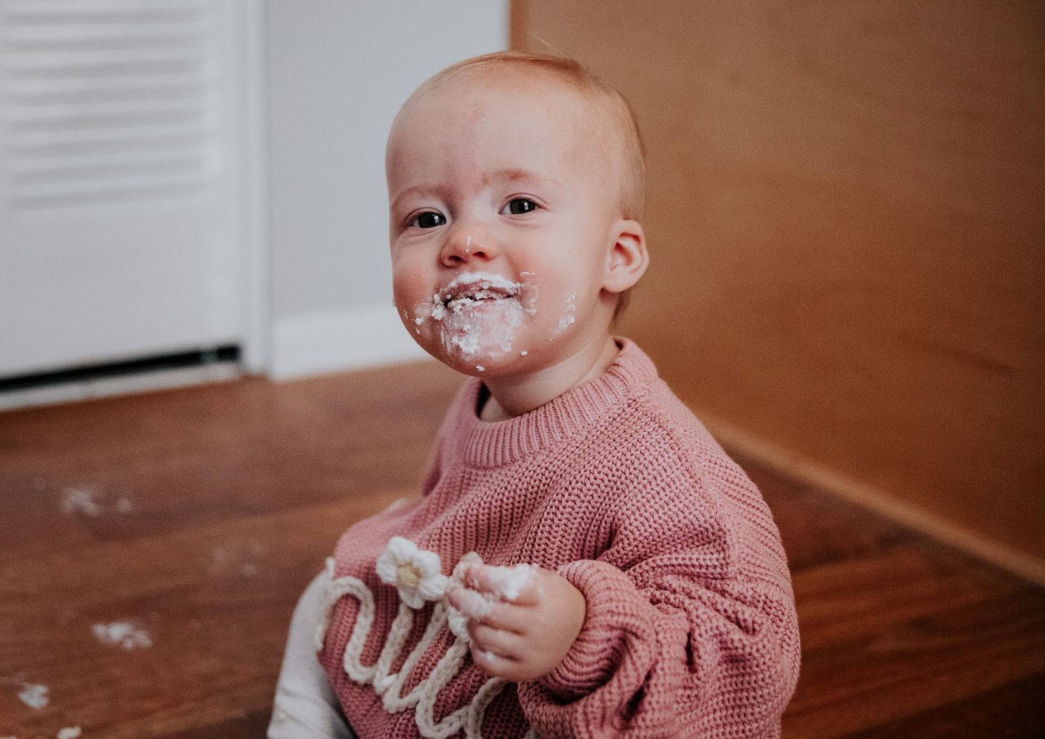 A messy cake smash photo with frosting on face and a pink knit sweater.
