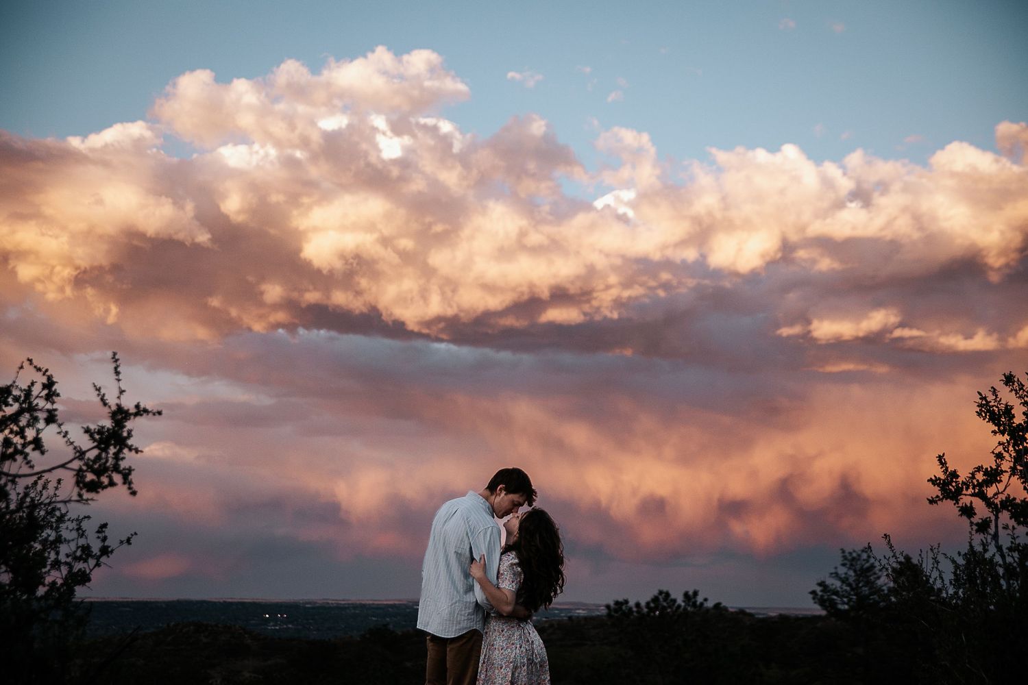 Silhouetted couple embraces under dramatic sunset clouds in mountain landscape.