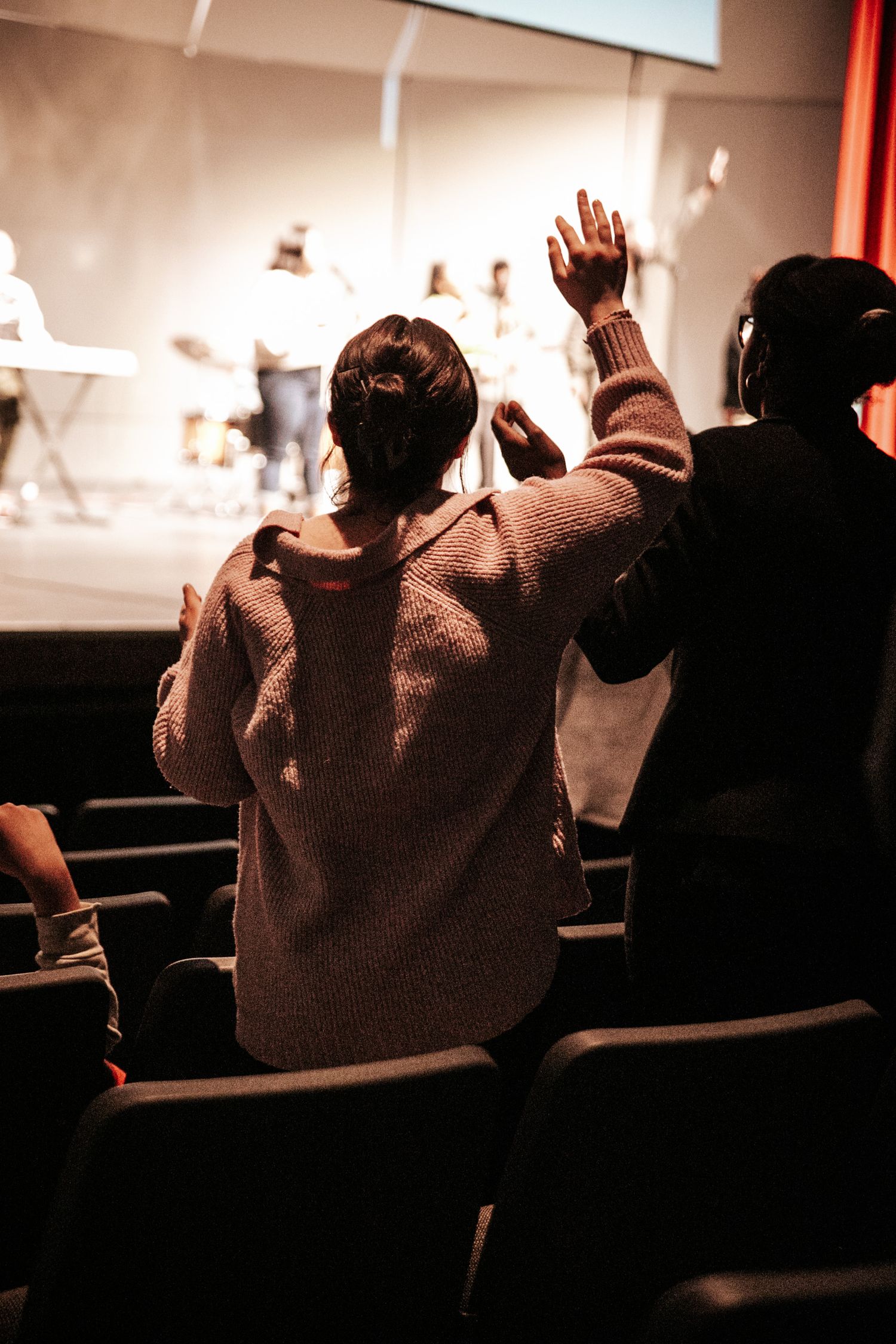 Person raising hand during worship service in dimly lit auditorium.