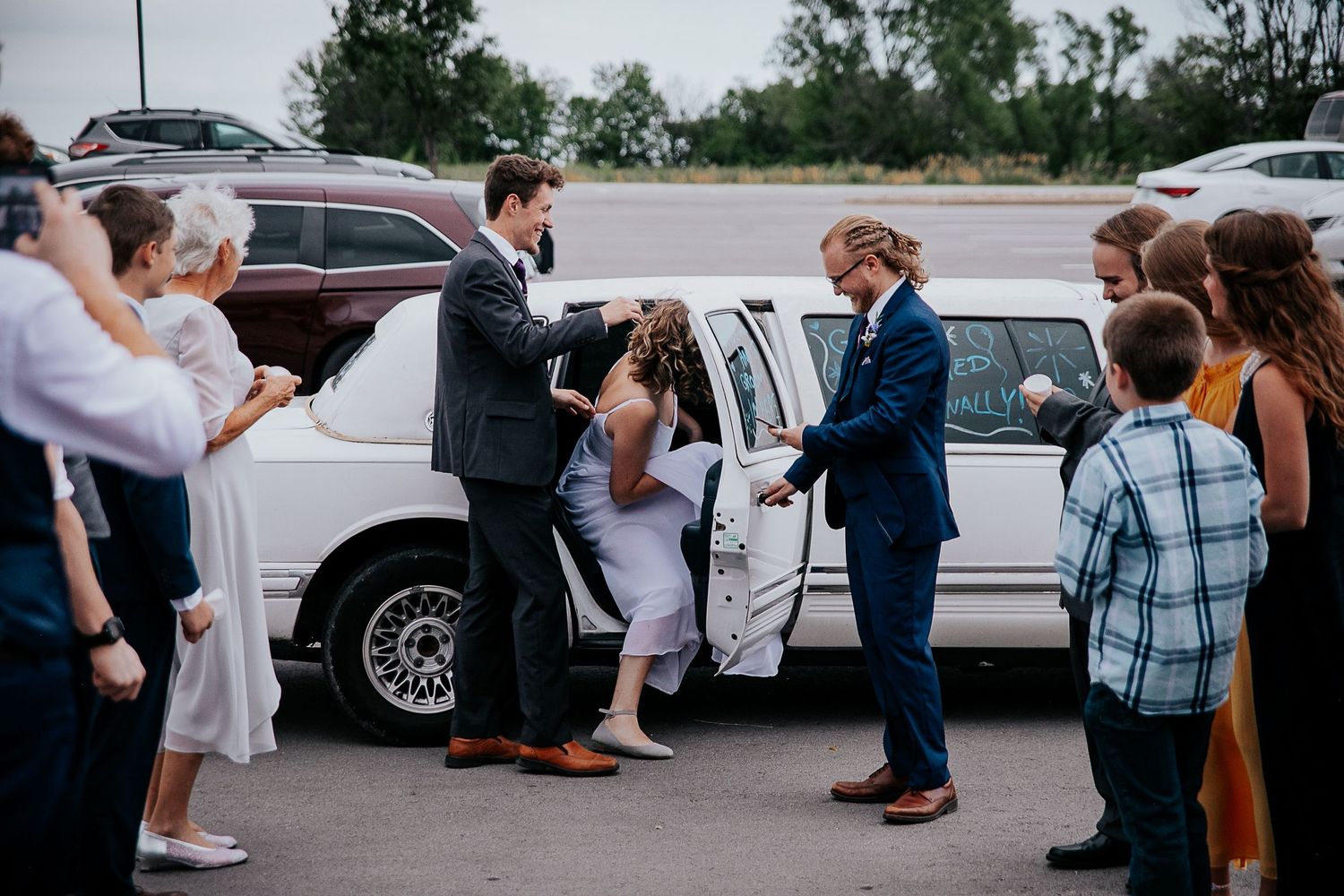 Wedding party members help the bride into a white limousine after the celebration.