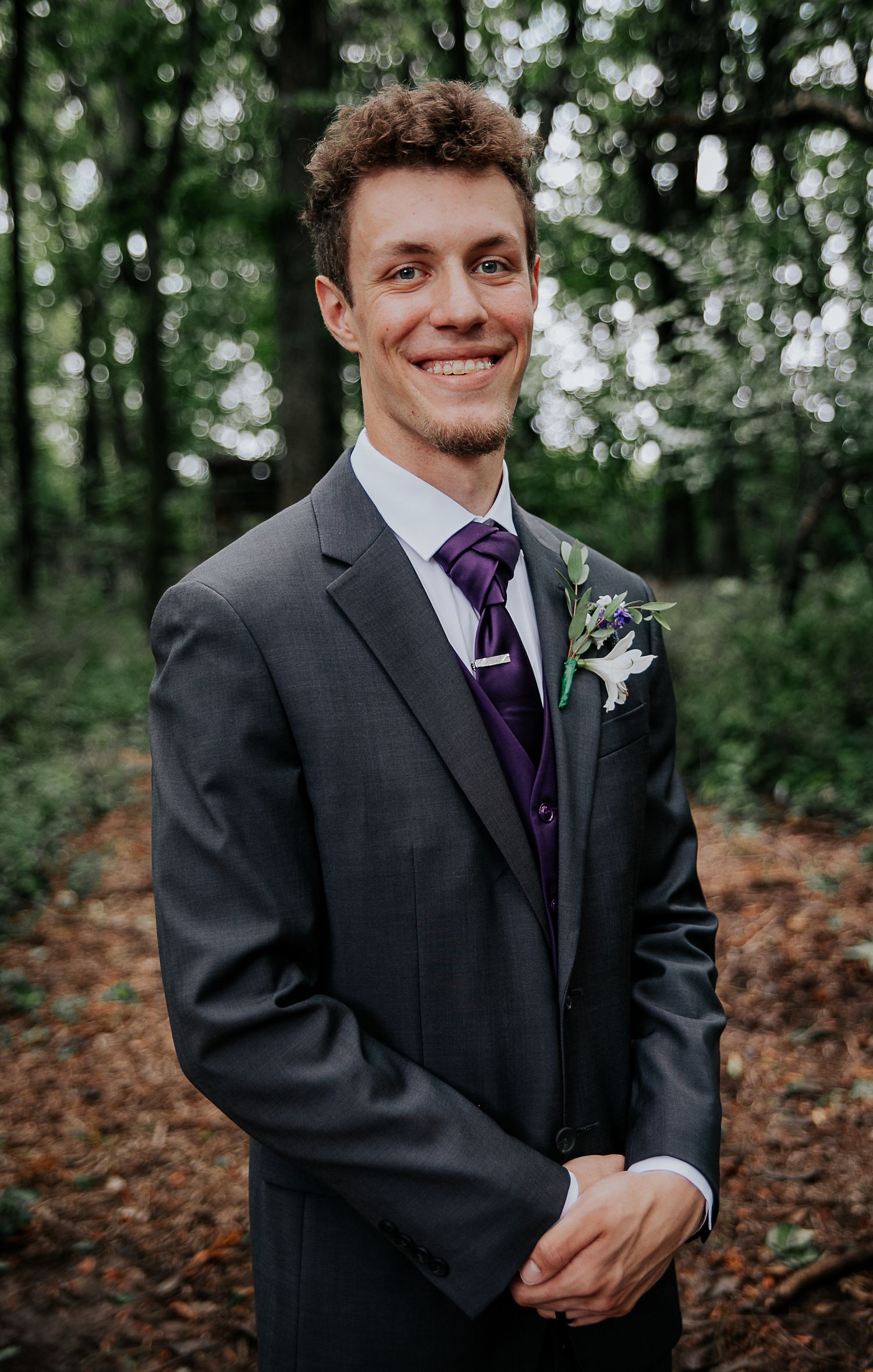 Groom in dark suit with purple tie and boutonniere smiles in woodland setting.