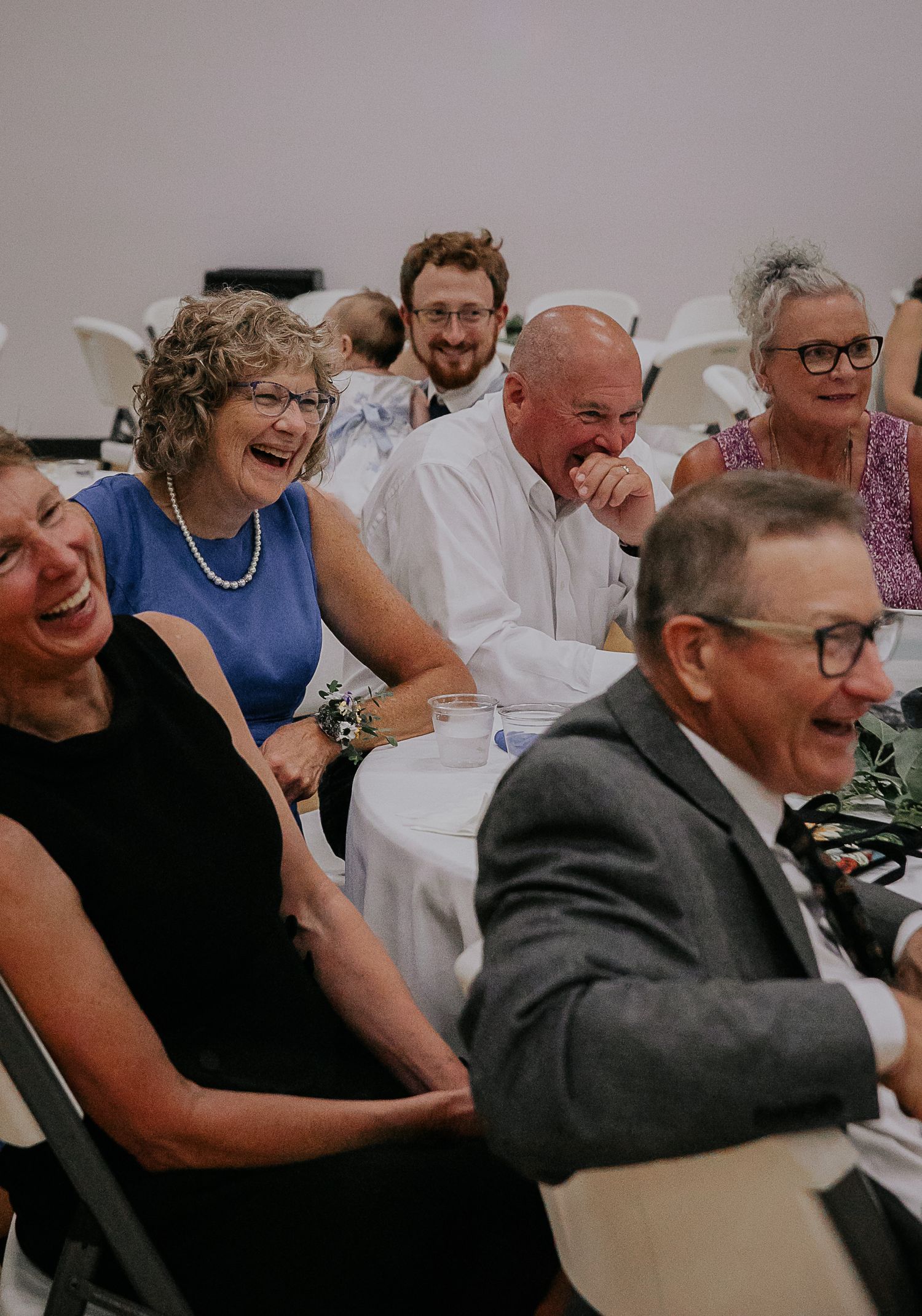 Wedding guests laugh joyfully together during reception celebration in white event space.