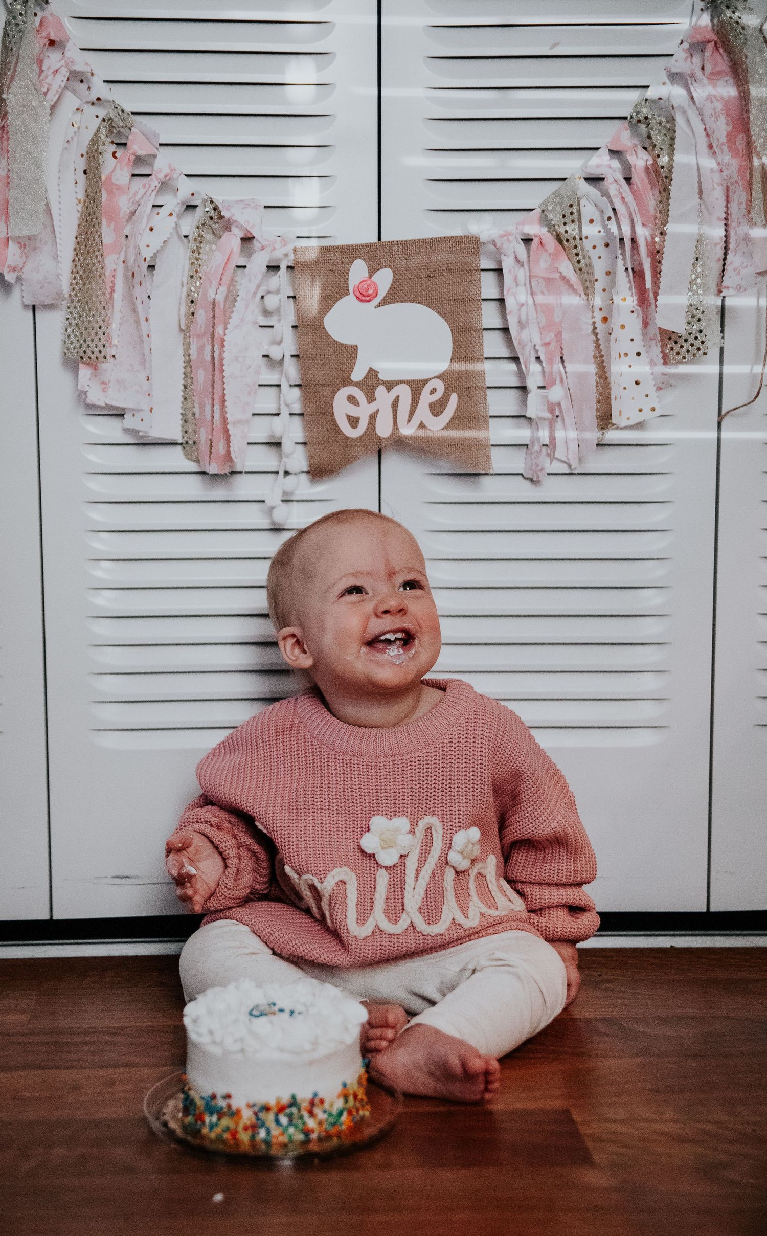 Smiling baby wearing pink sweater next to white frosted cake with sprinkles.