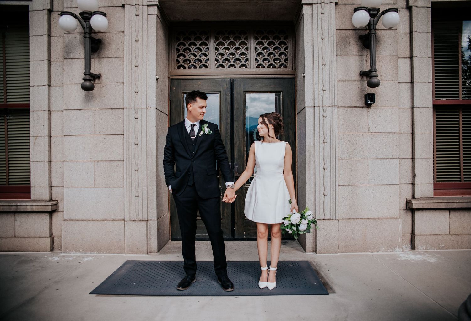 A couple in wedding attire stands on a stone staircase outside a historic building with ornate architecture.