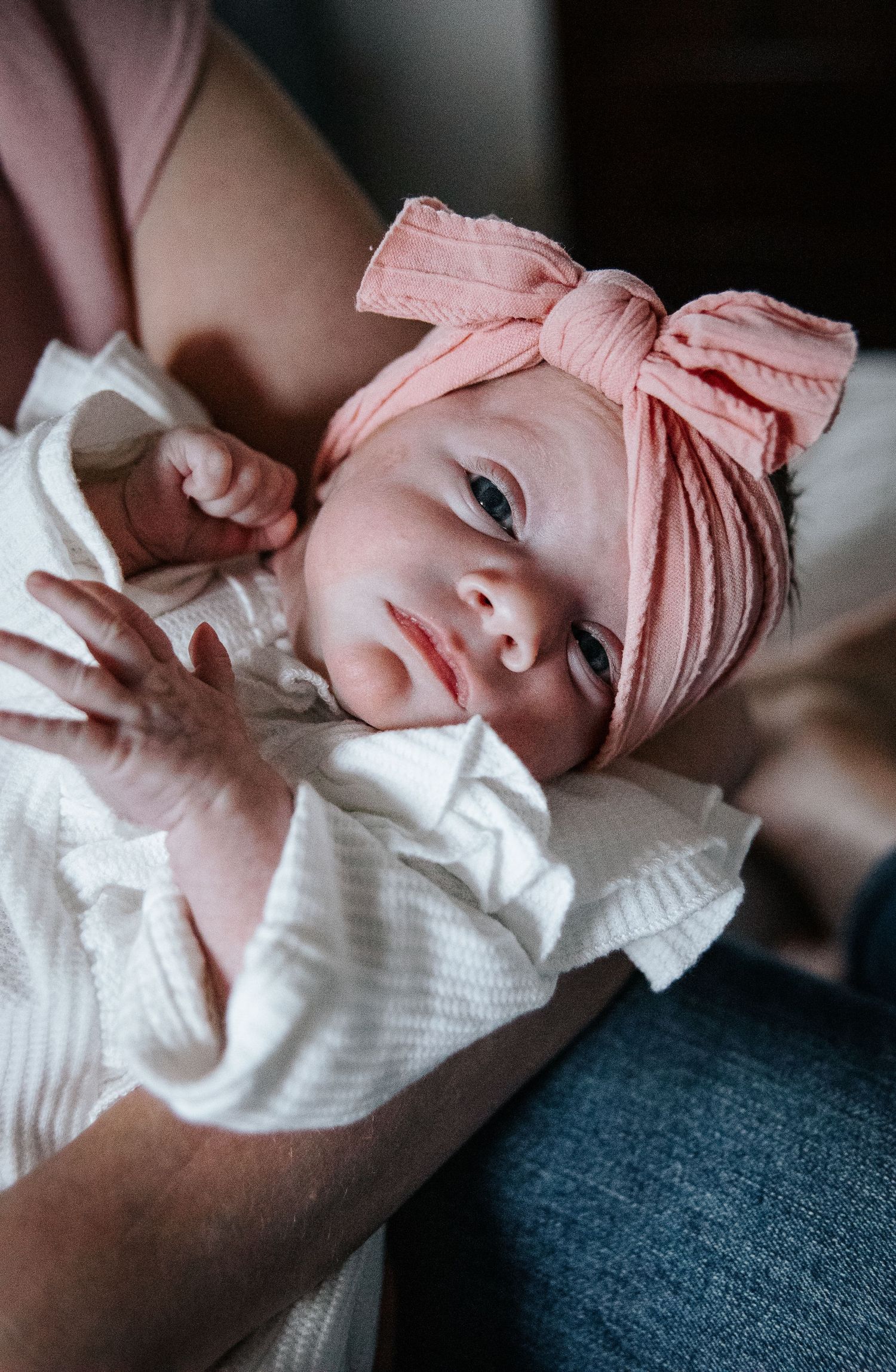 Baby wearing pink headwrap bow and white ruffled outfit rests while being held on denim lap.