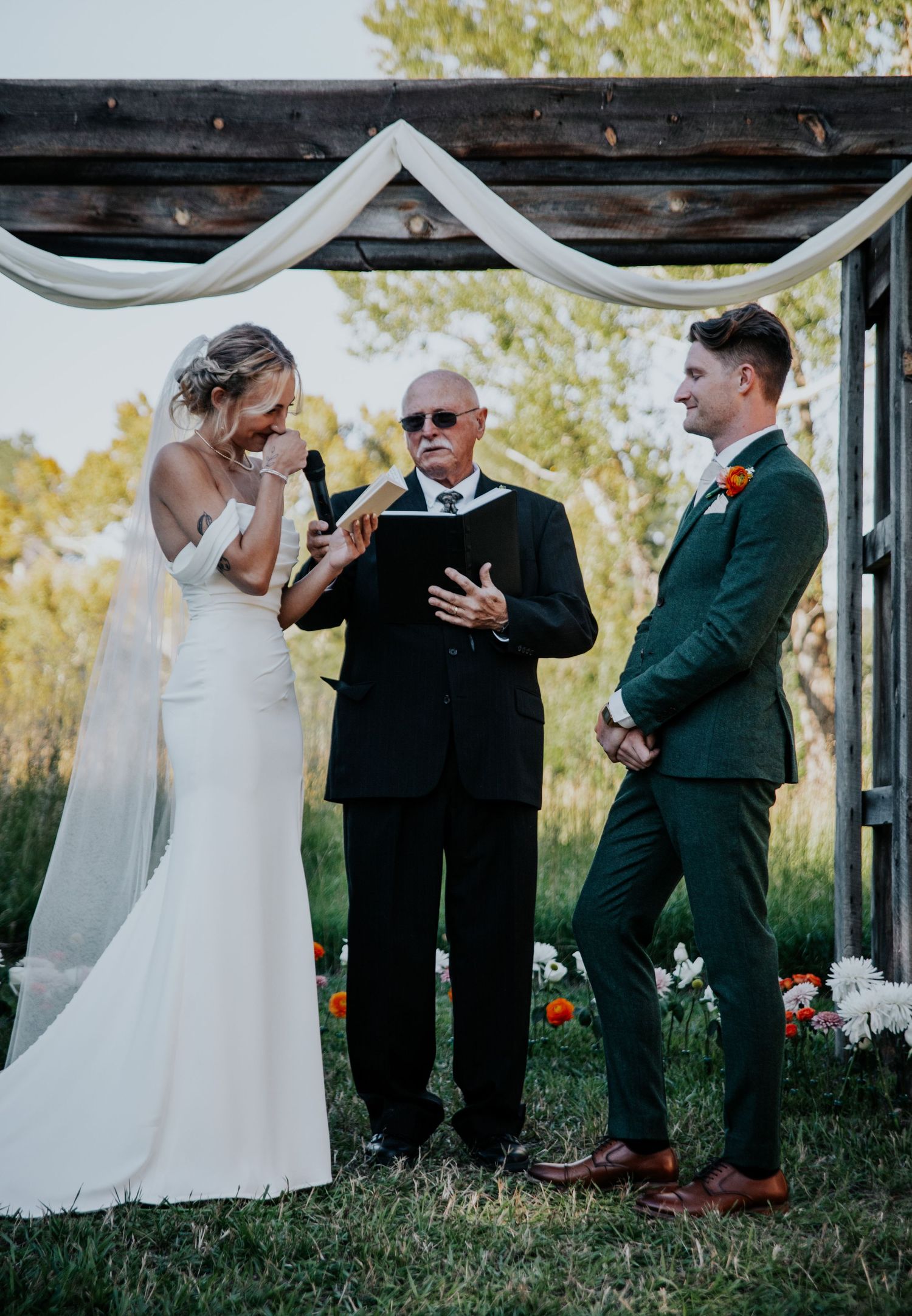 A wedding ceremony takes place under a rustic wooden arch decorated with white fabric and flowers.