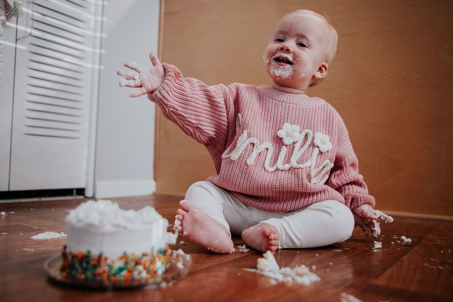 Baby in pink sweater sits on floor with birthday cake and smeared frosting, expressing pure joy.