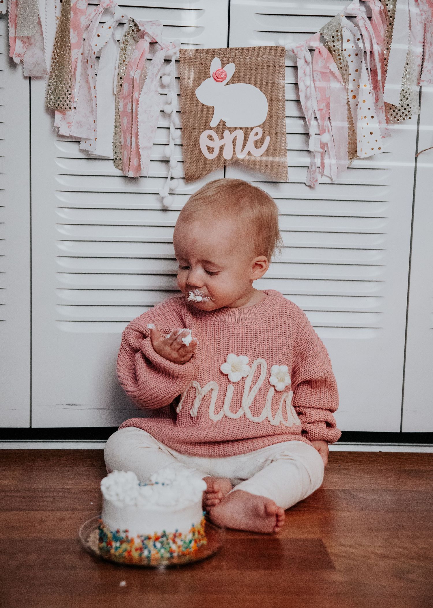 Baby in pink knit sweater laughing while playing with birthday cake on wooden floor.