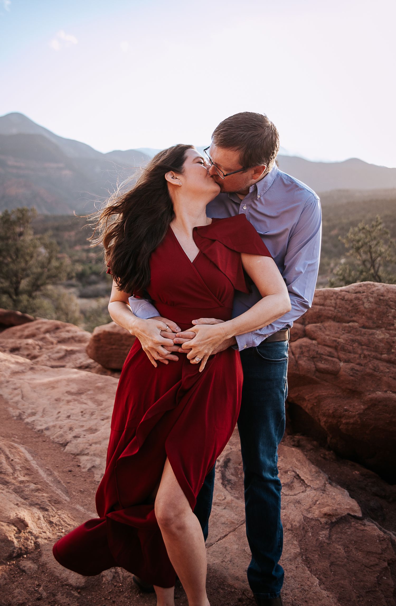 A couple in red and blue embrace on red rocks with mountains in the background.
