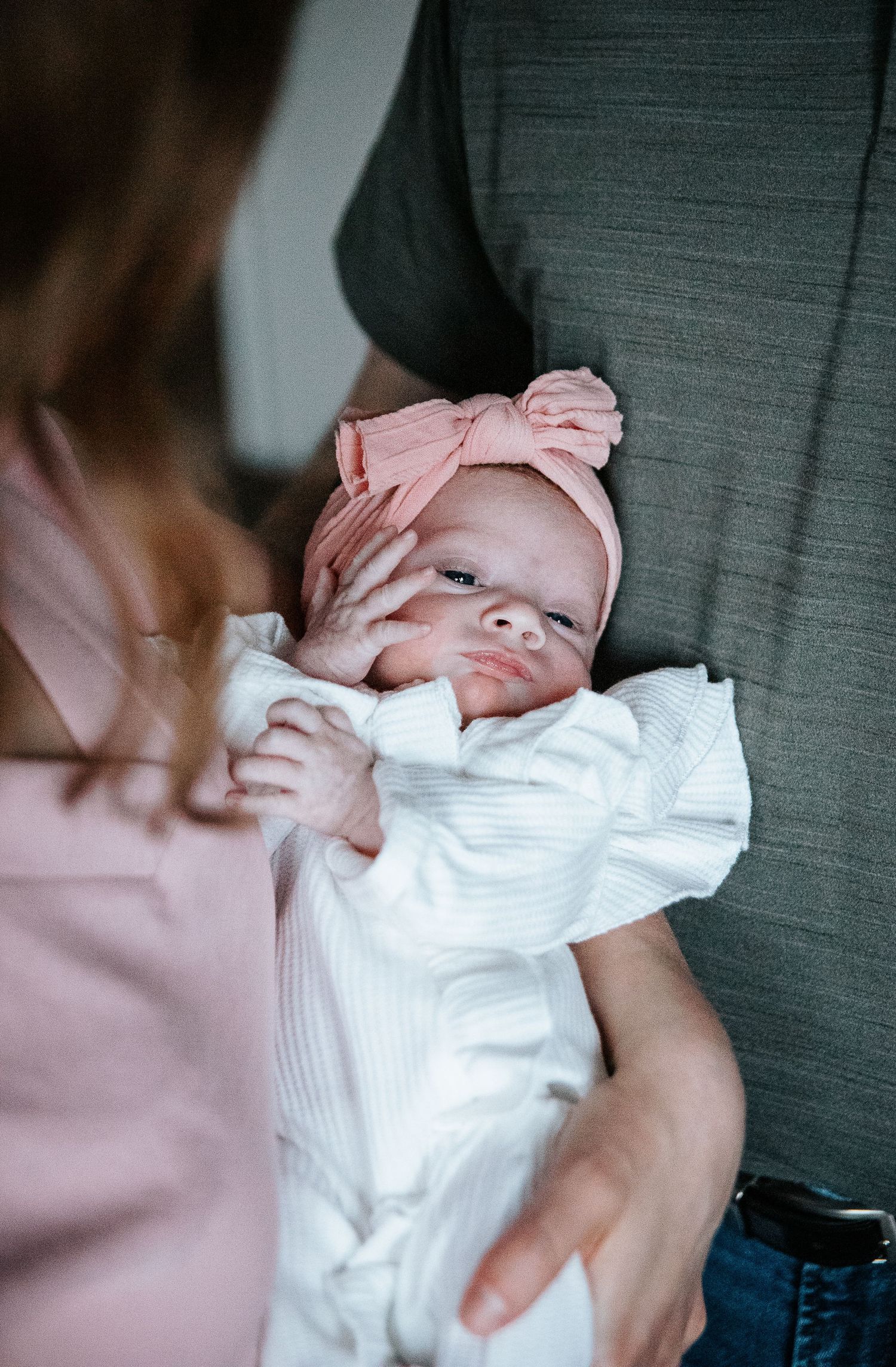 A newborn wearing a white outfit and pink bow headband being cradled in someone's arms.
