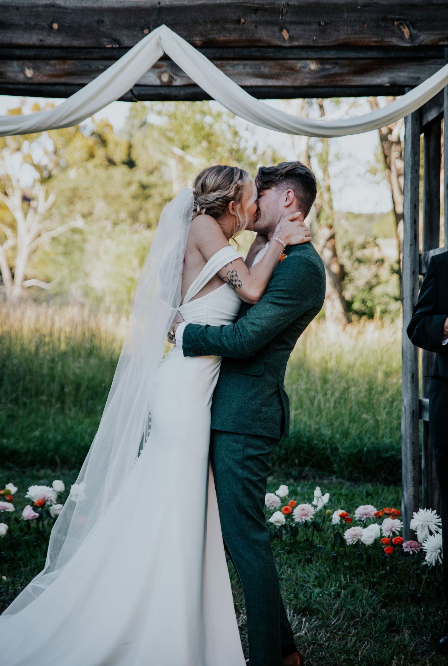 A romantic moment captured under a wooden arch decorated with white fabric and flowers at an outdoor wedding.