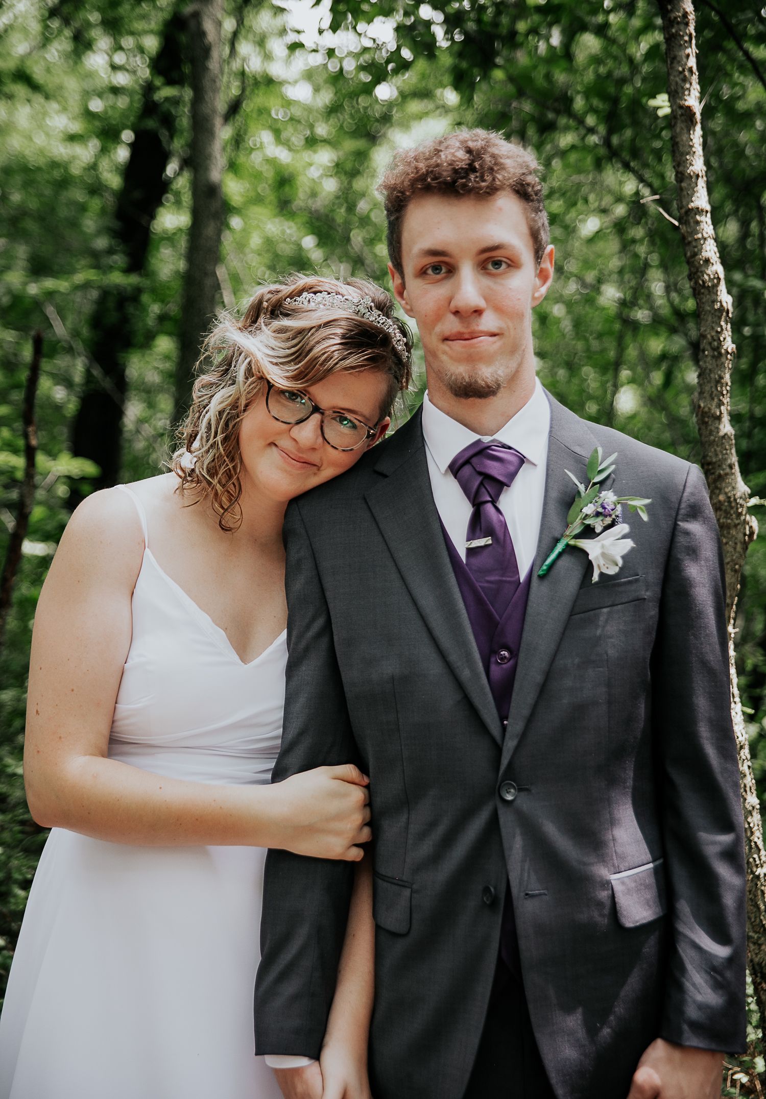Couple poses together in woods wearing wedding attire against natural backdrop.