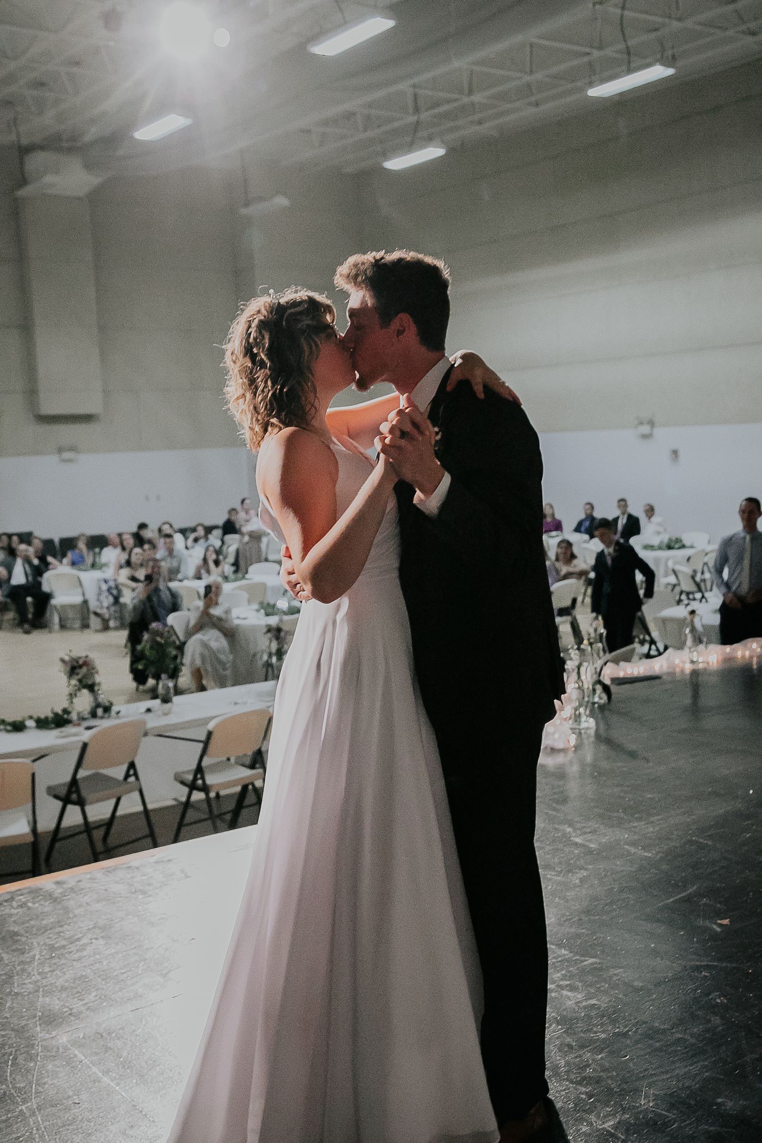 Bride and groom share romantic dance in industrial reception venue with guests in background.