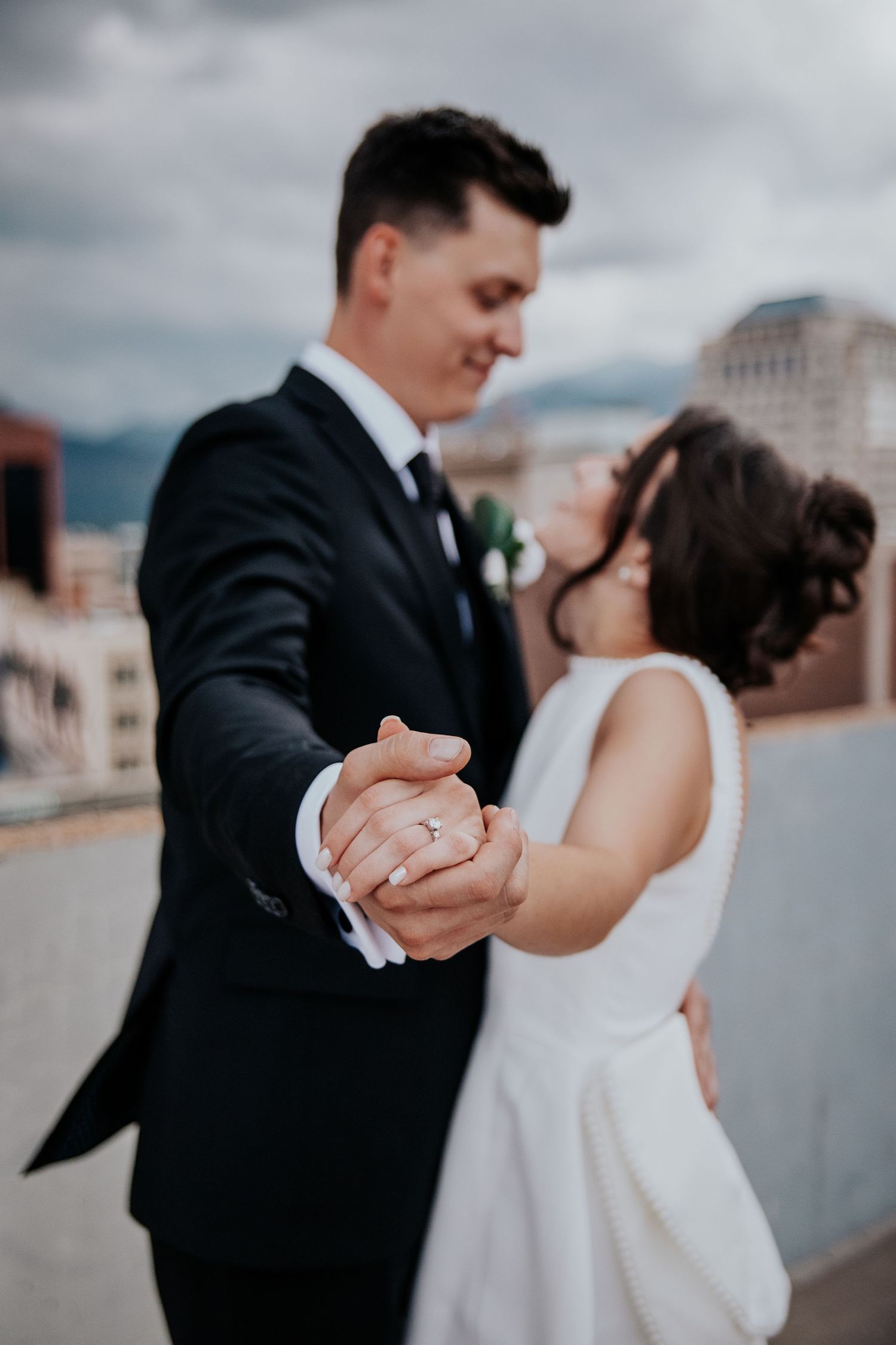 Newlywed couple dancing together on a rooftop with cloudy skies and city buildings in the background.