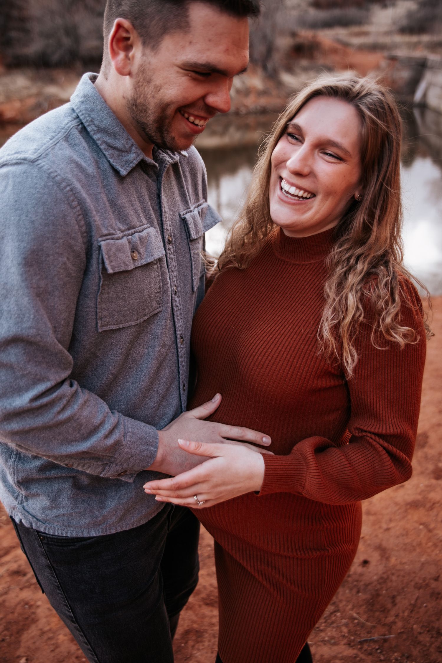 A joyful maternity moment captured outdoors with a couple in casual attire sharing laughter against a desert backdrop.
