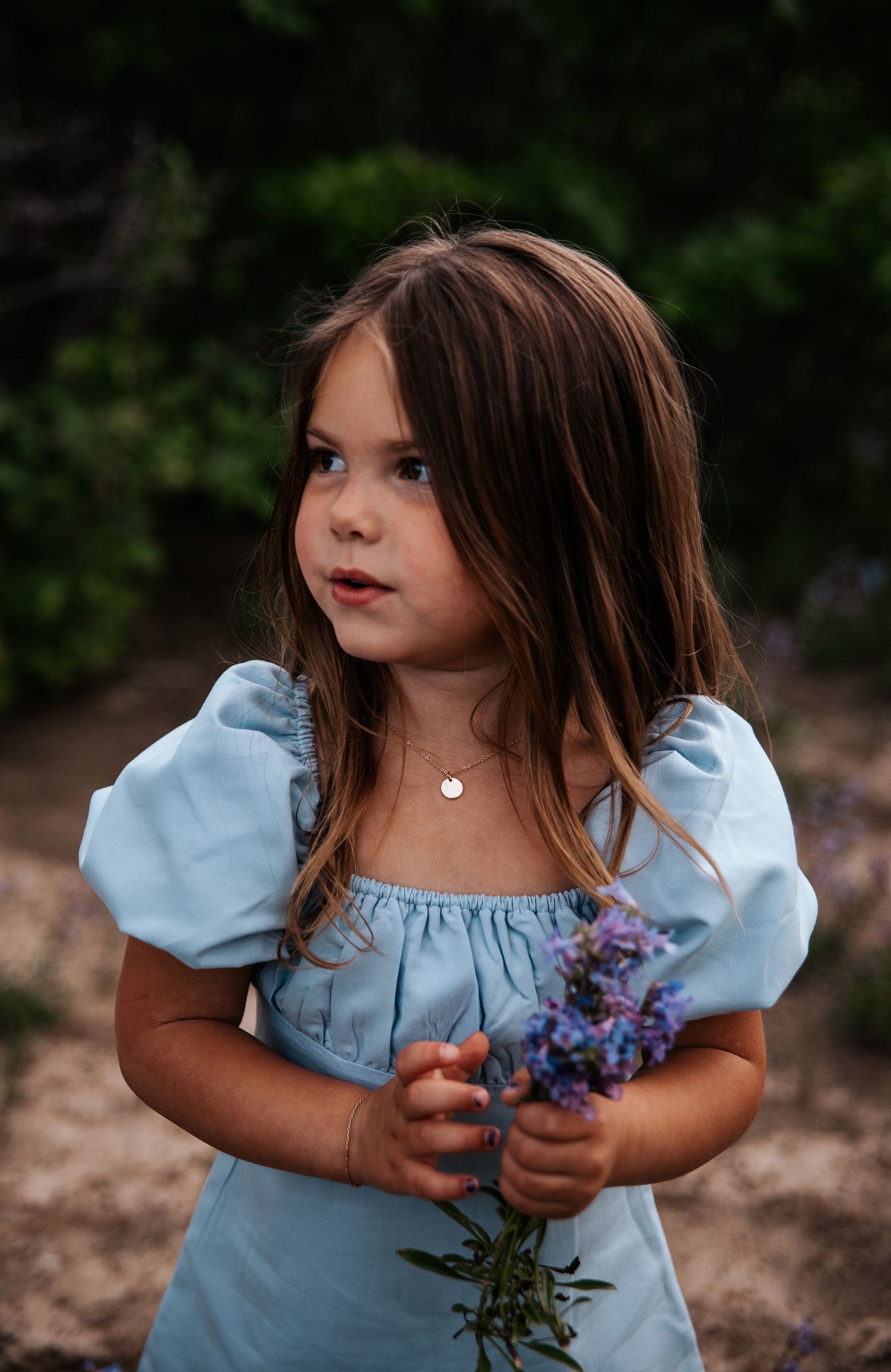 Little girl in light blue dress holds purple flowers while posing in nature at Ute Valley Park in Colorado Springs