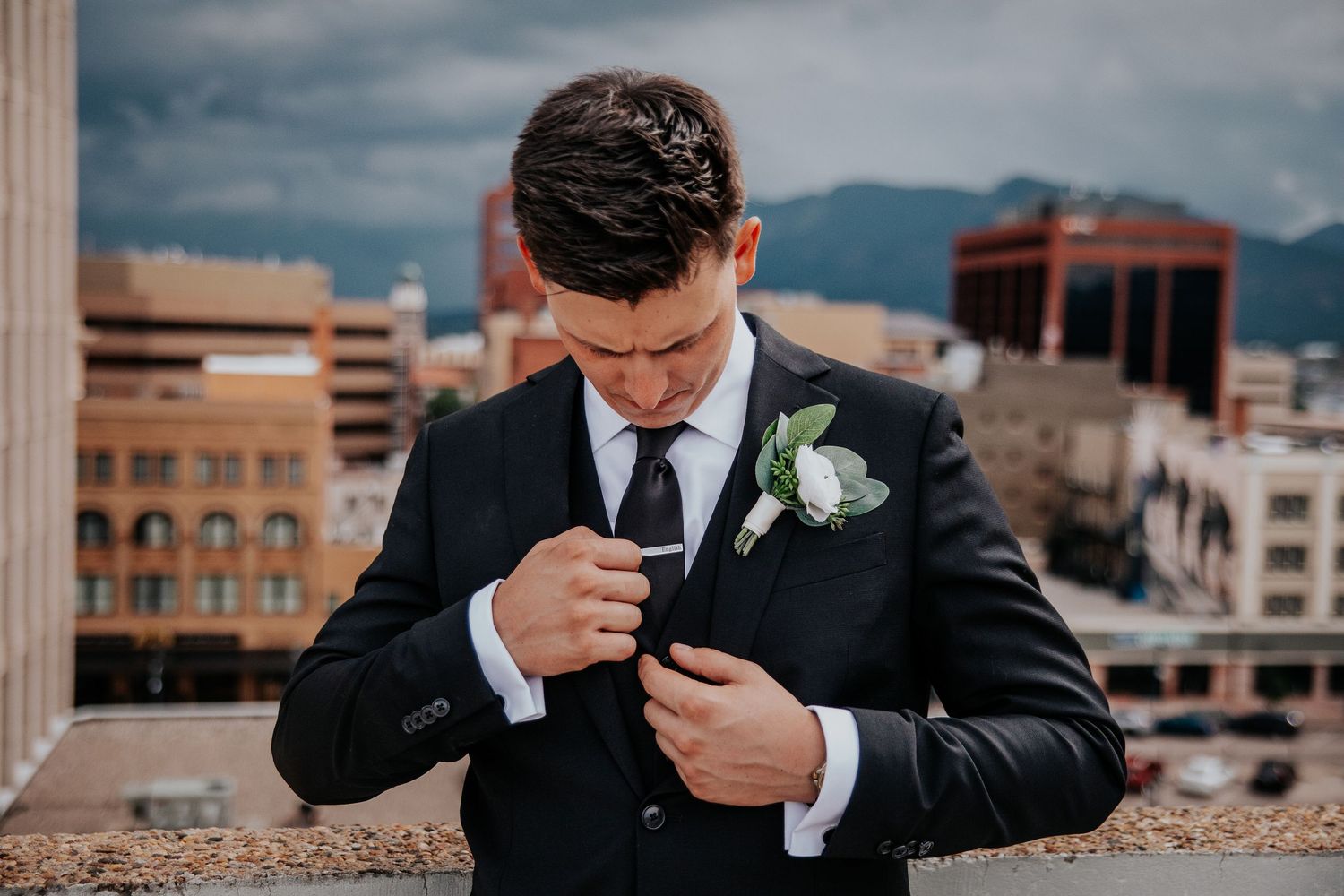 Groom in black suit wipes tears during emotional wedding moment against urban backdrop.