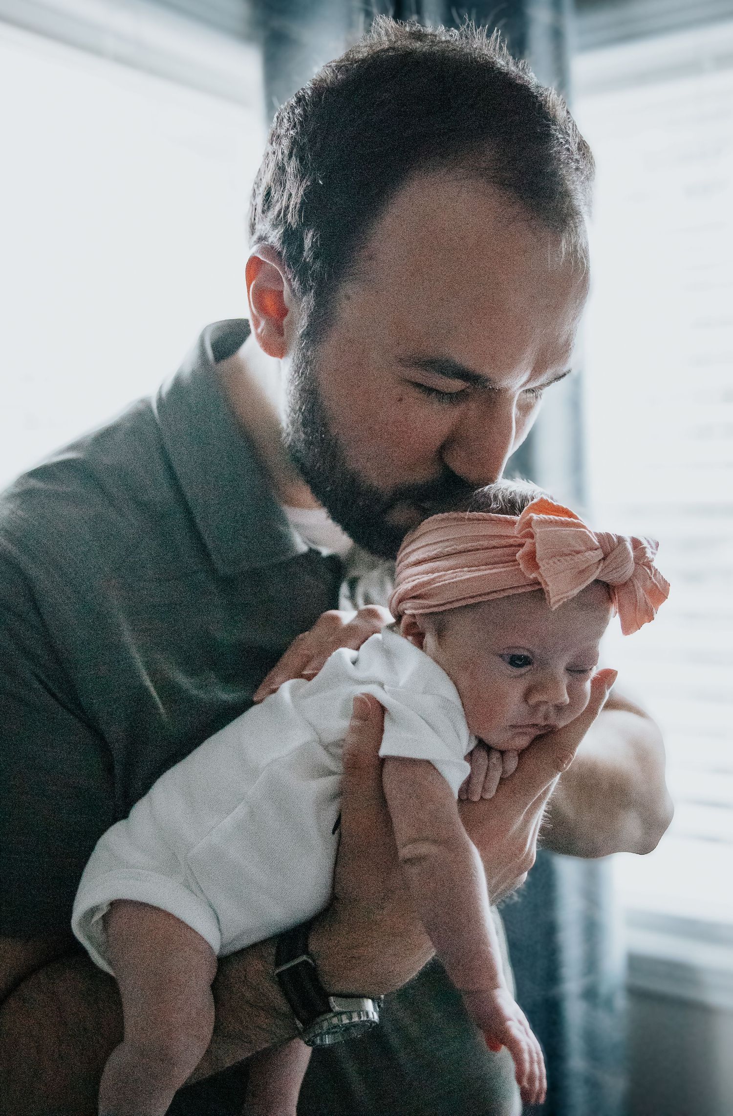 A tender moment as a bearded parent in a gray sweater holds and kisses their infant wearing white clothing.