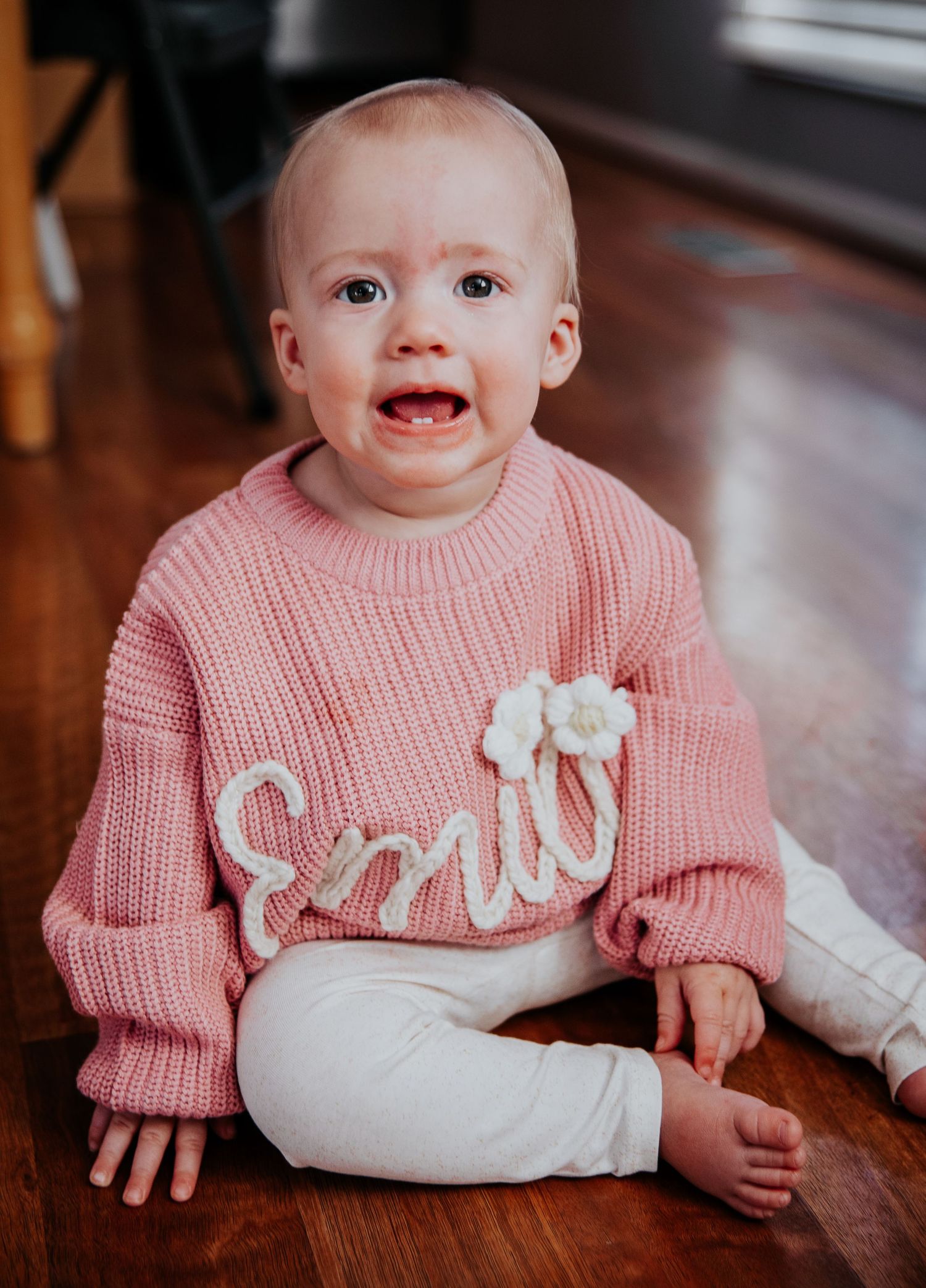 Baby in pink knit sweater sitting on hardwood floor expressing emotion.