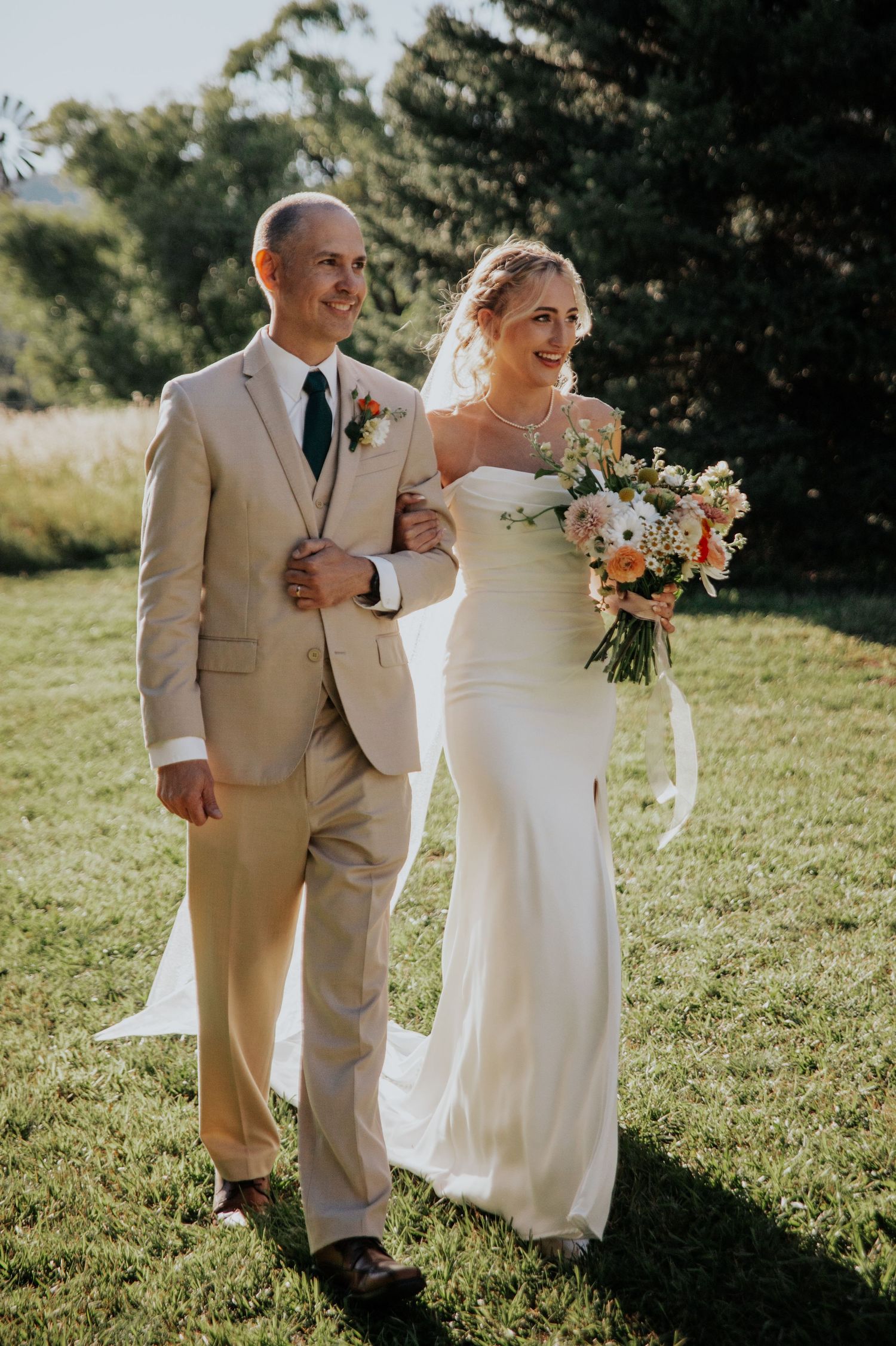 A couple walks arm-in-arm on their wedding day, with the bride in white and groom in beige against a sunny outdoor backdrop.