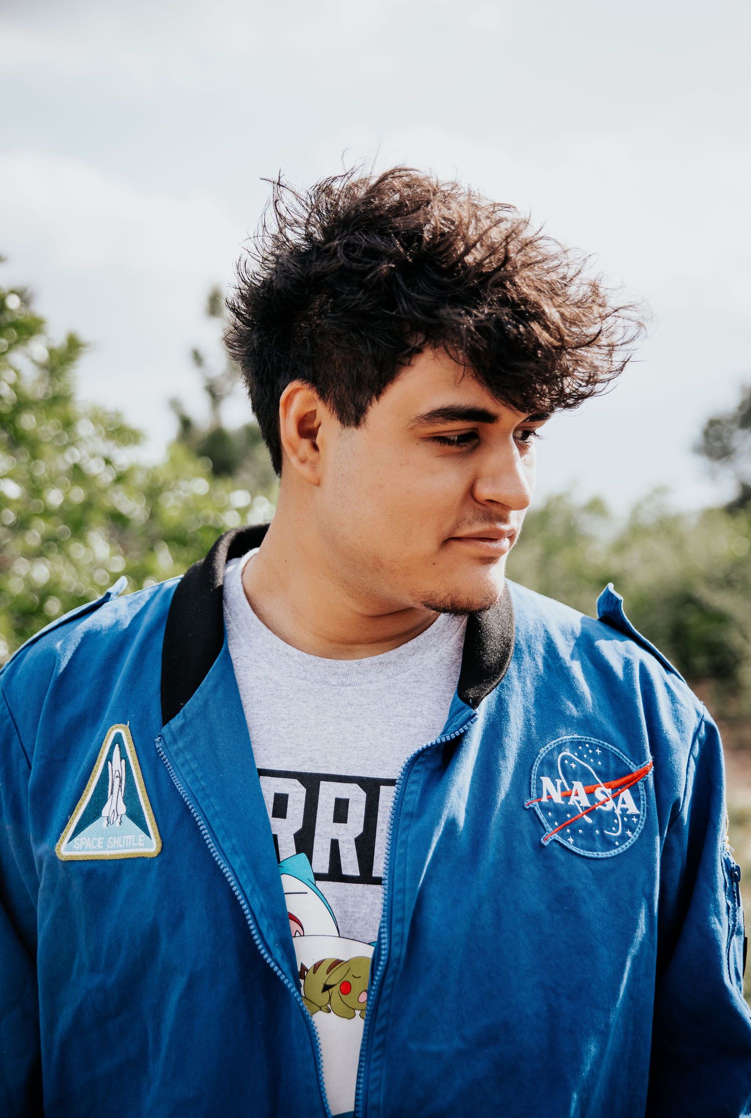High school senior wearing NASA uniform and white t-shirt stands against cloudy sky backdrop at Palmer Park in Colorado Springs