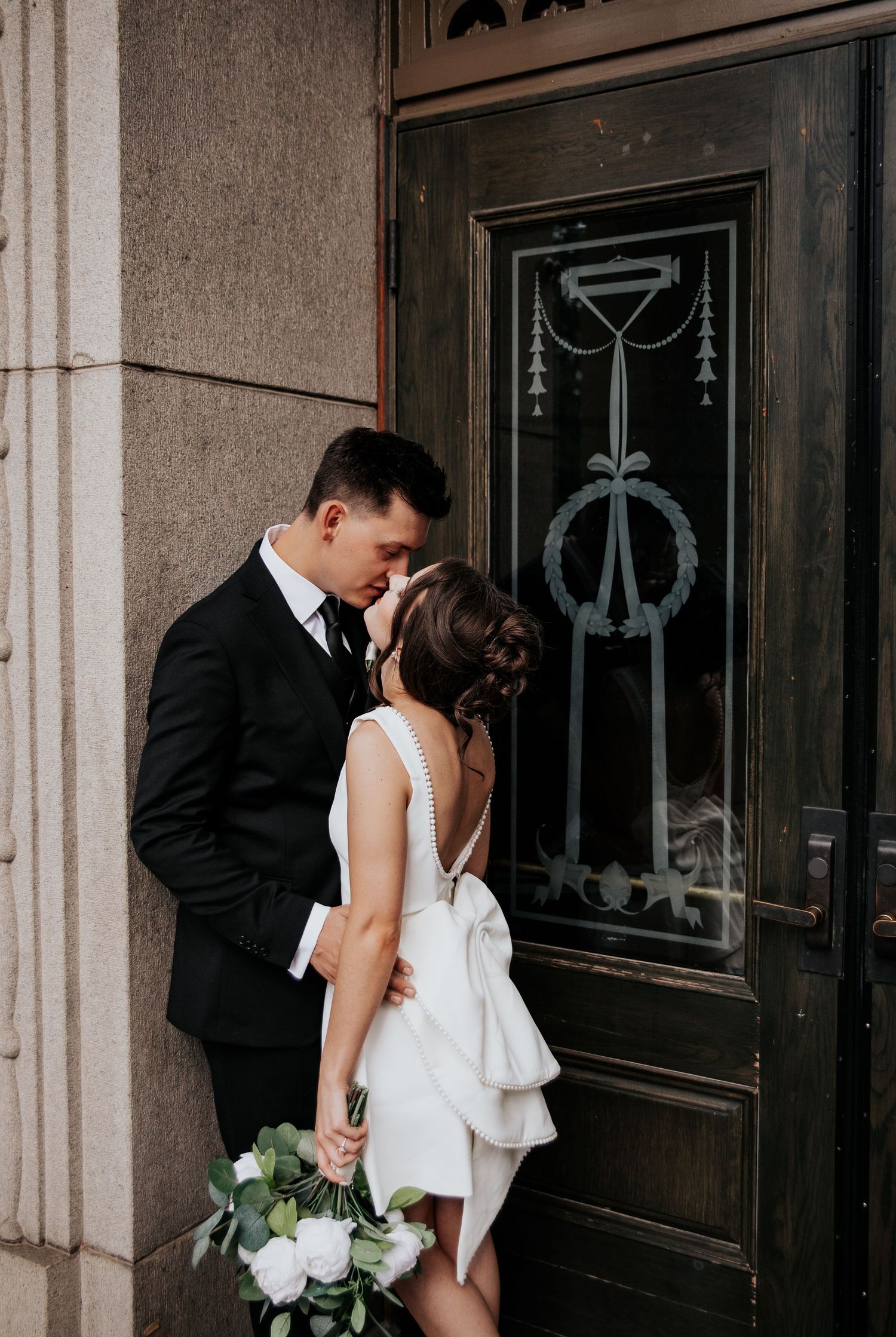 A romantic wedding couple shares an intimate moment in front of ornate black doors at an elegant venue.