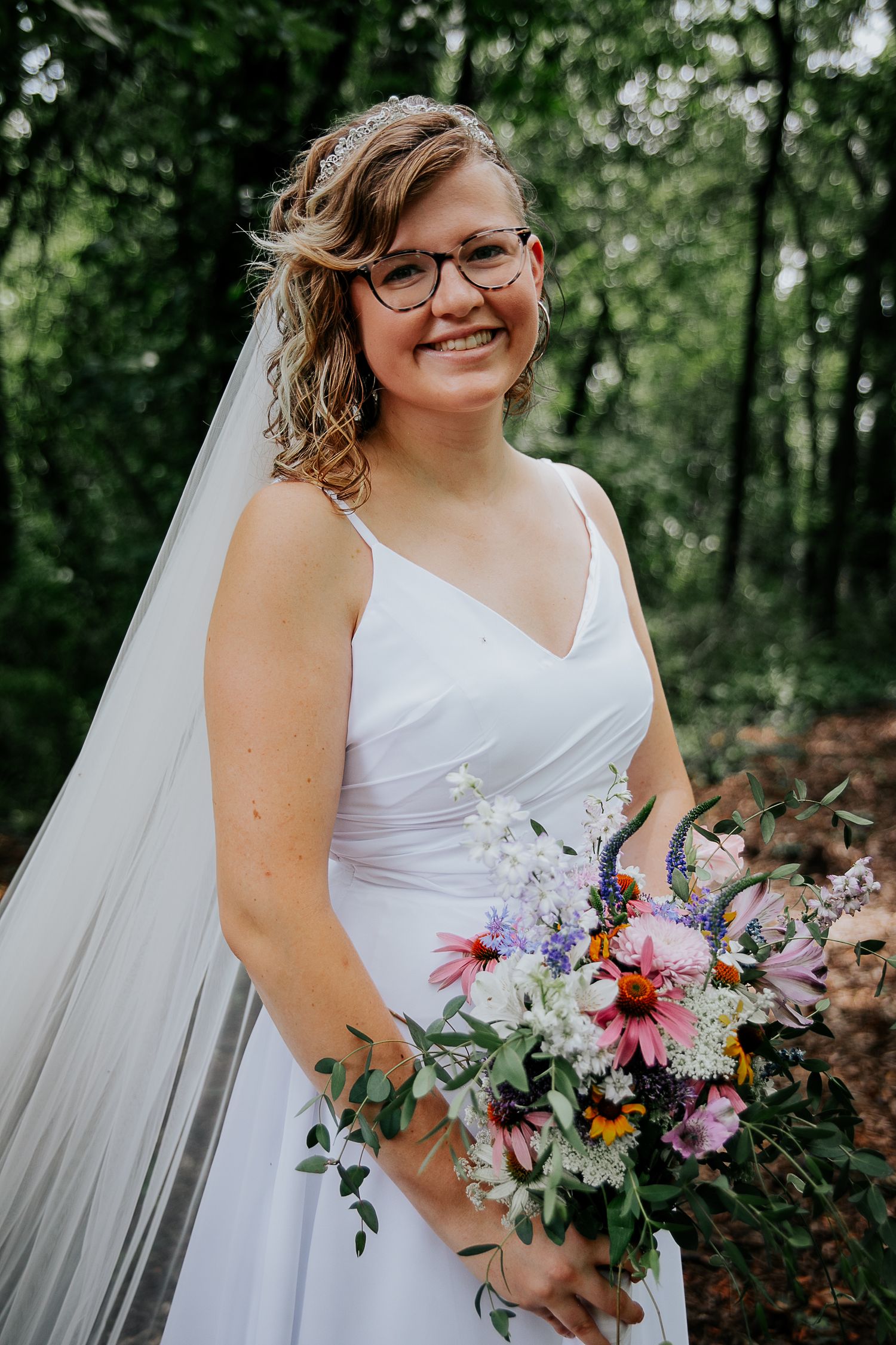Bride in white dress and glasses holds colorful wedding bouquet in wooded setting.