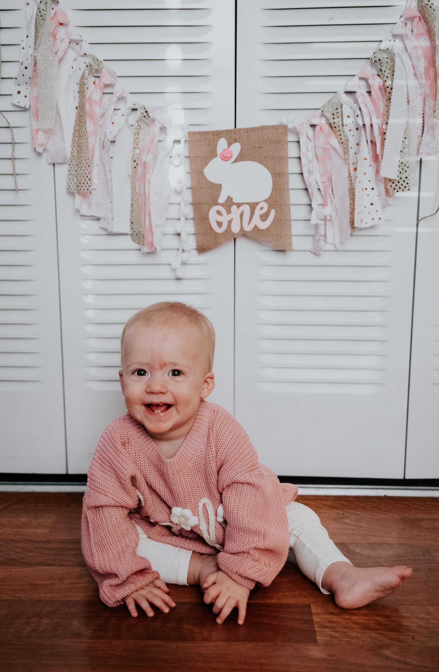 Baby in pink sweater sits by decorated wall with one sign and banners.