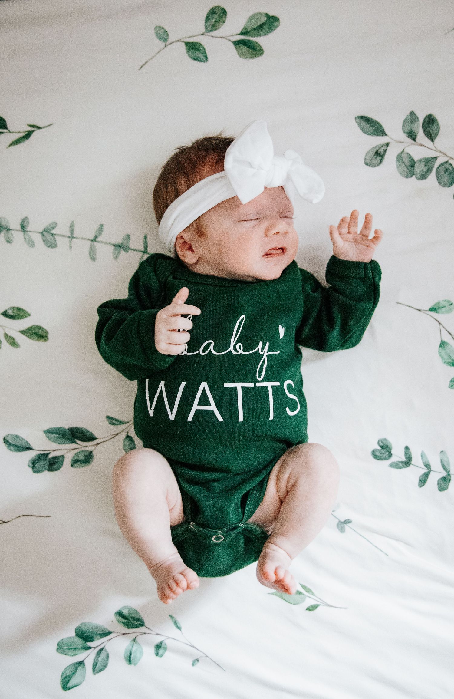 Baby in dark green sweater lying on white bedding with eucalyptus leaf decorations on the wall.