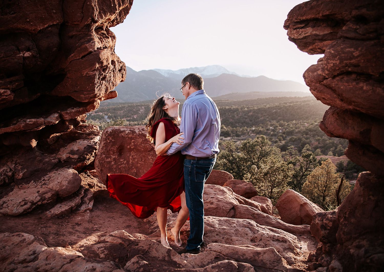 A couple stands together on red rock formations overlooking a mountain valley.