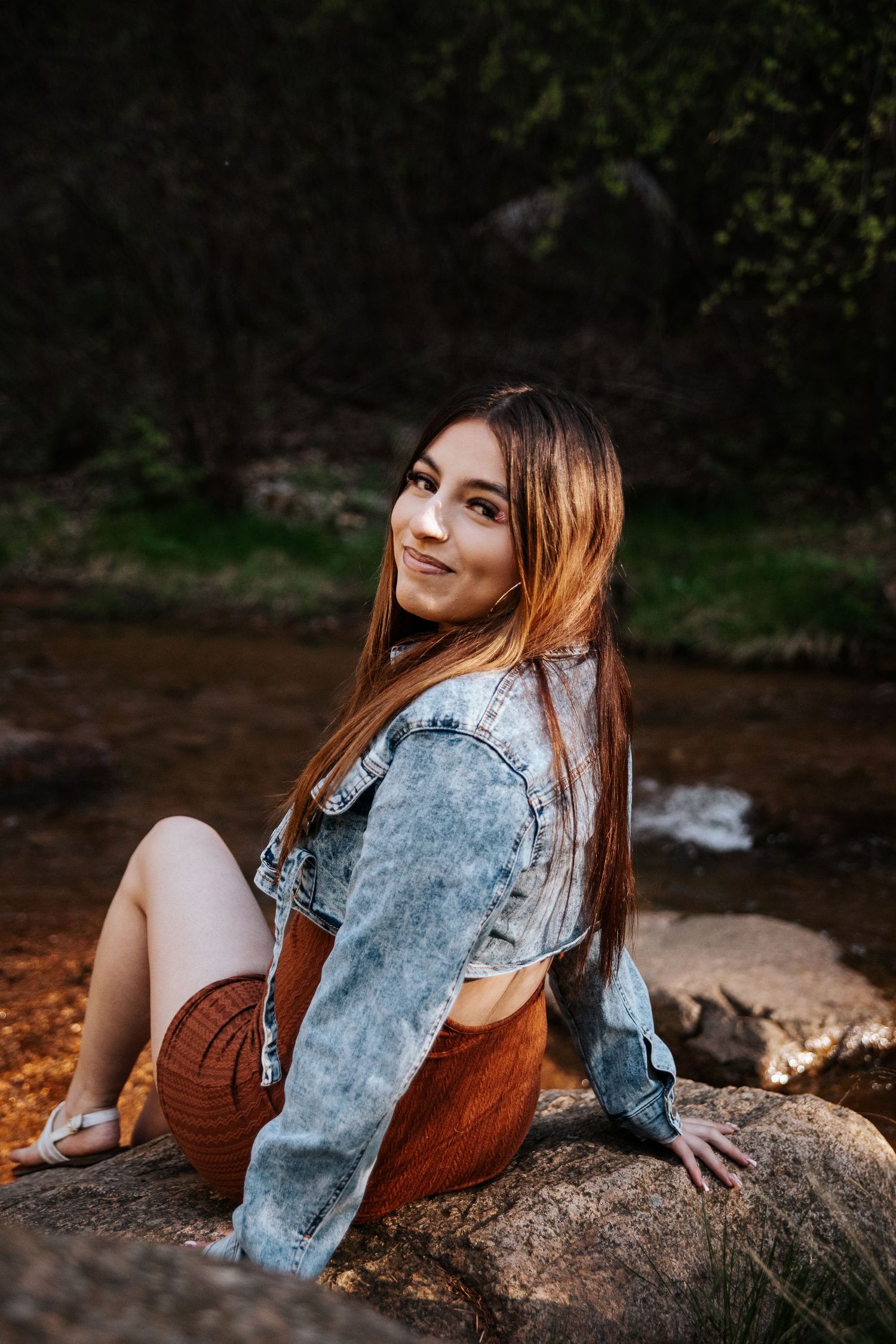 Portrait sitting on rocks by stream wearing denim jacket and brown dress in natural setting.
