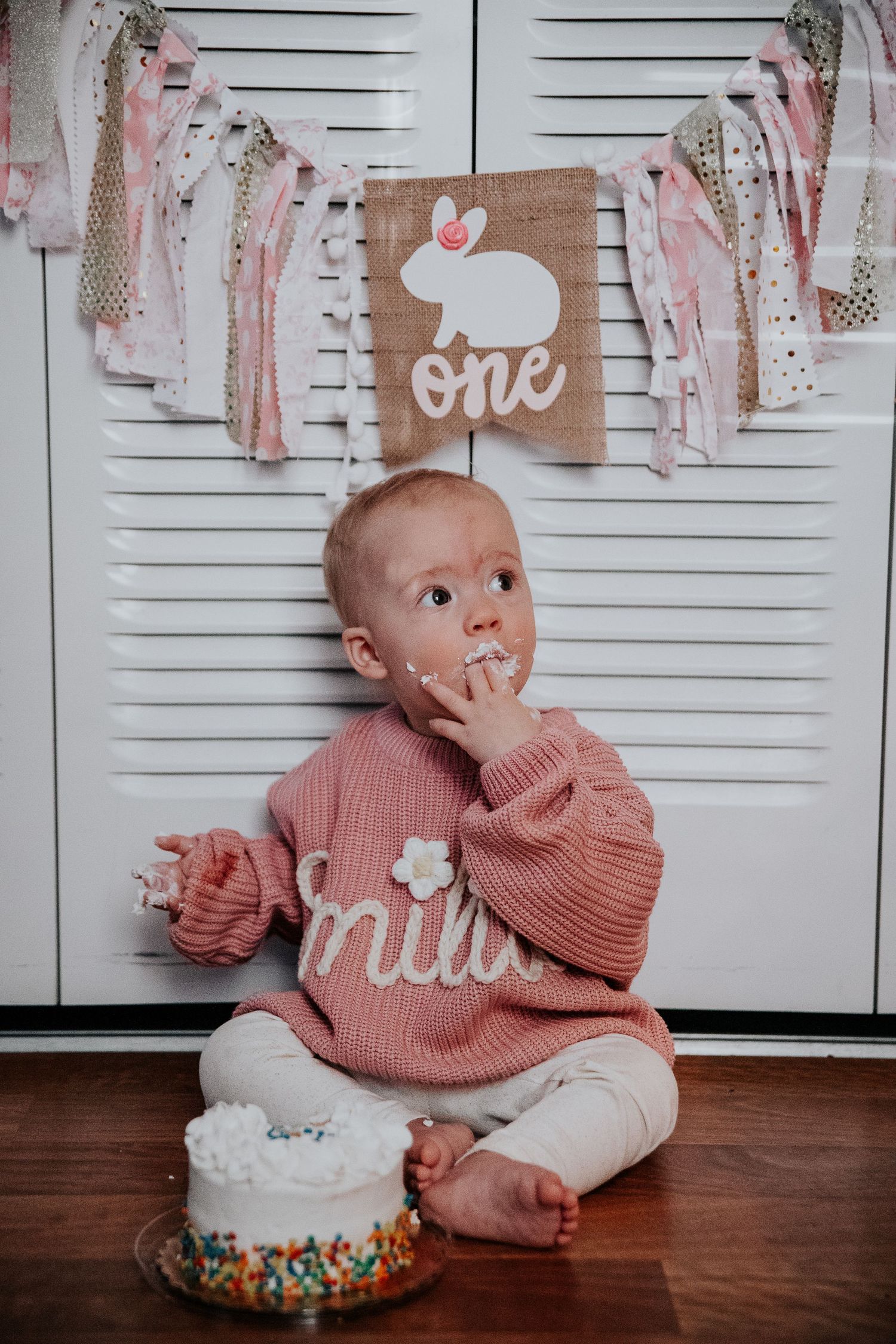 Baby in pink sweater looking up during first birthday cake smash session.