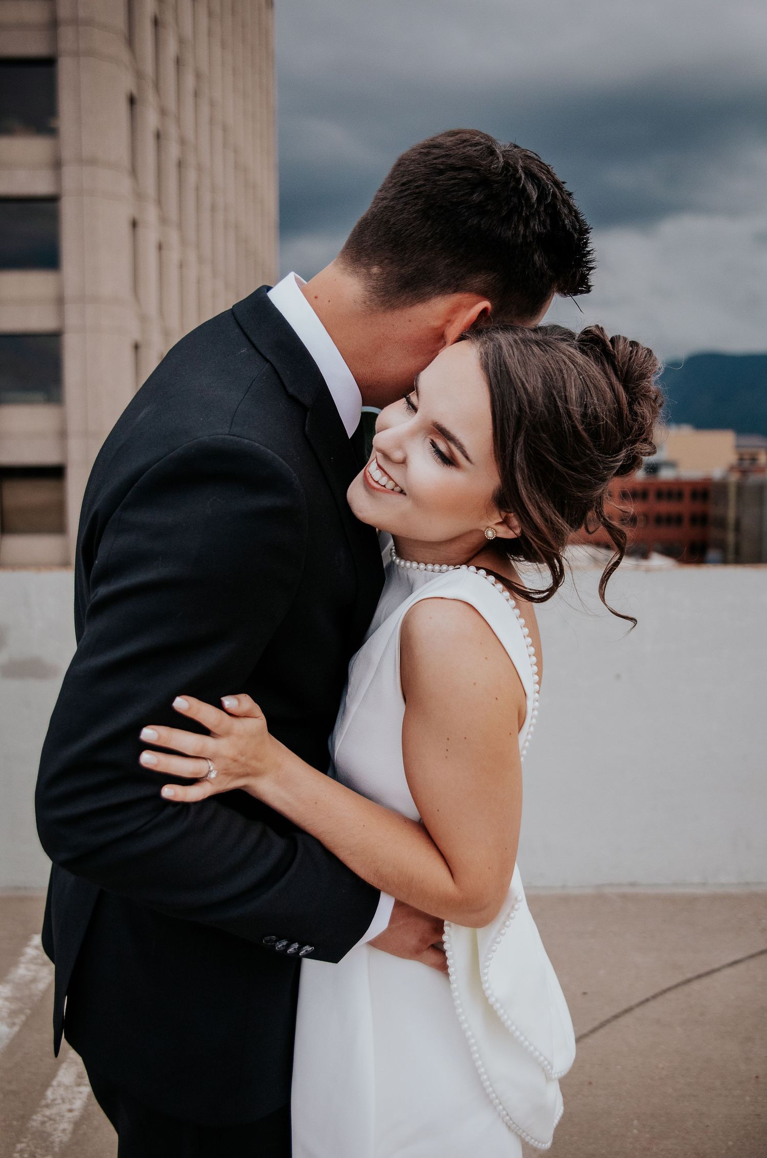 Intimate moment between newlyweds embracing on city rooftop under moody sky.