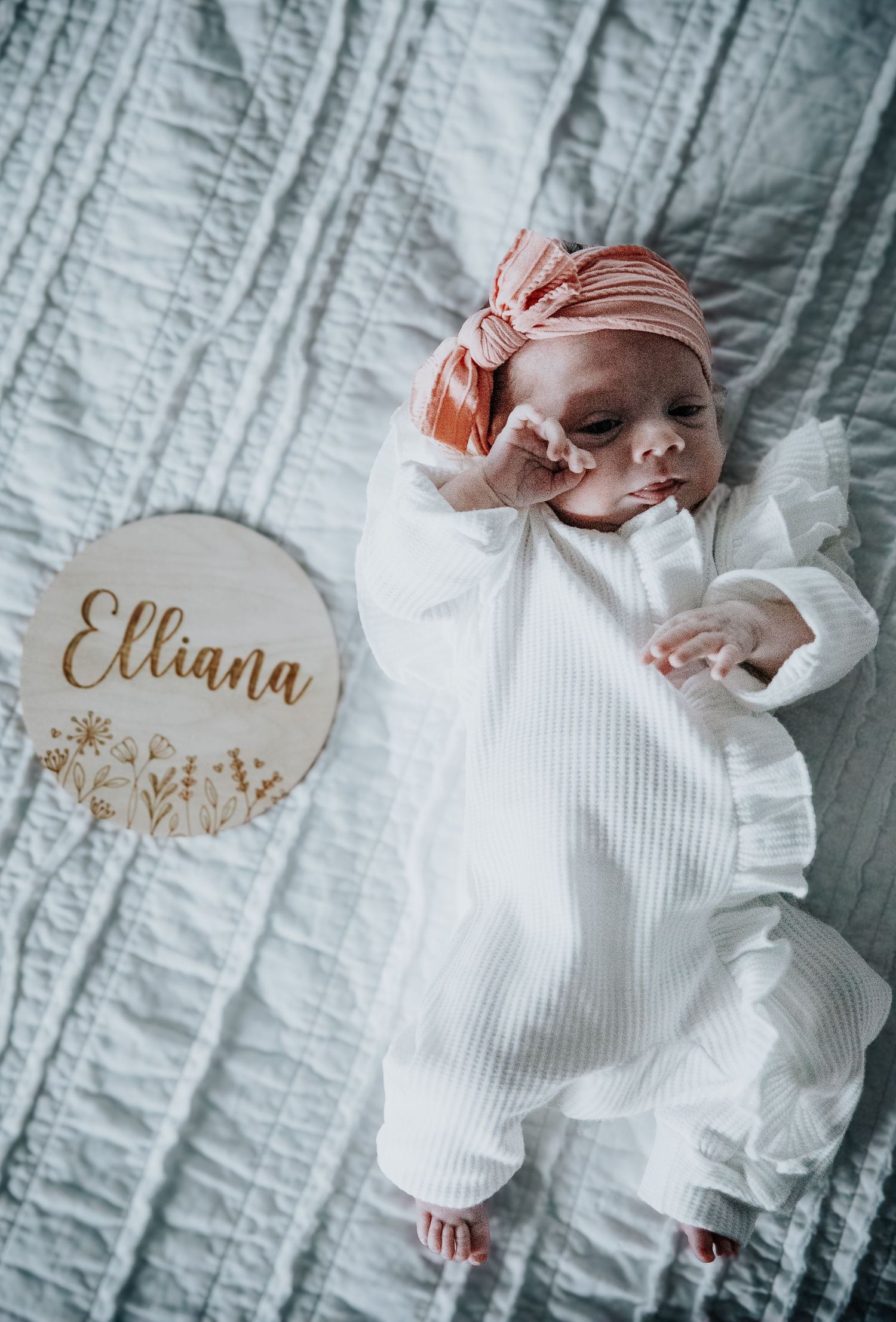 A newborn baby in white clothing lies on a textured white blanket with personalized name decorations.