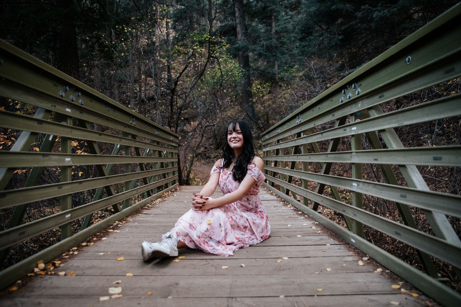 A floral pink dress flows across a wooden bridge surrounded by autumn foliage.