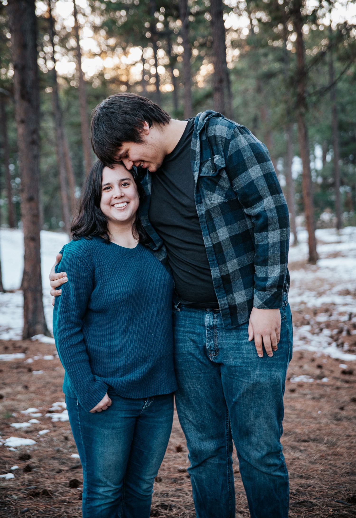 Loving couple embracing in snowy forest setting wearing casual winter clothing.