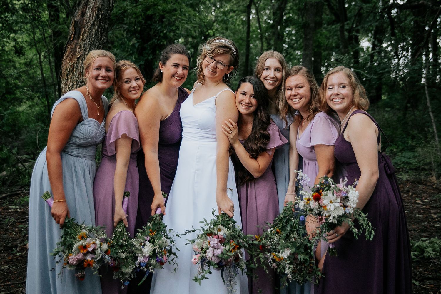 Wedding party in navy suits and purple dresses poses outdoors for a formal group photo.