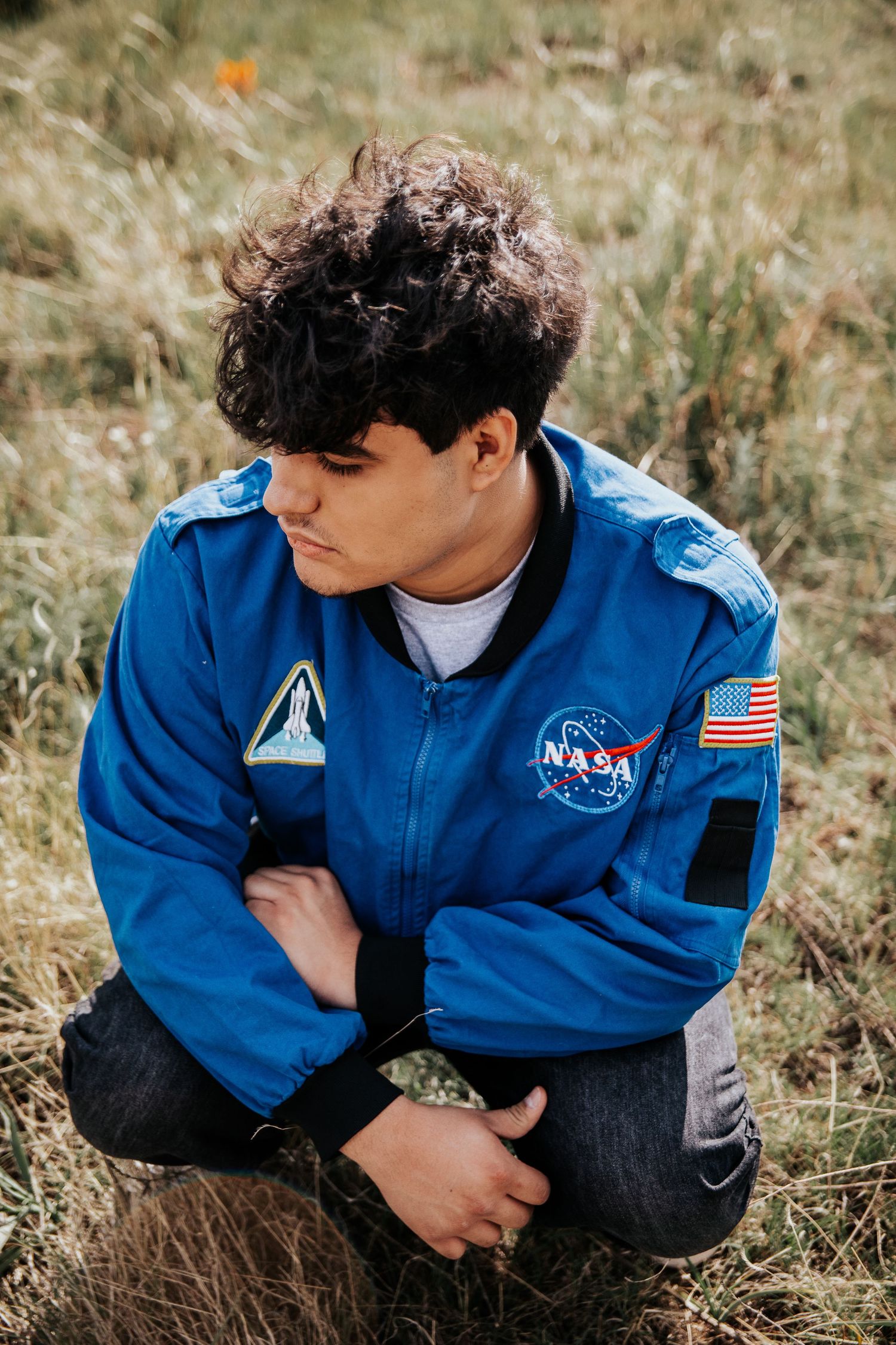 High school senior in blue NASA uniform sitting thoughtfully in wild grass field at Palmer Park in Colorado Springs