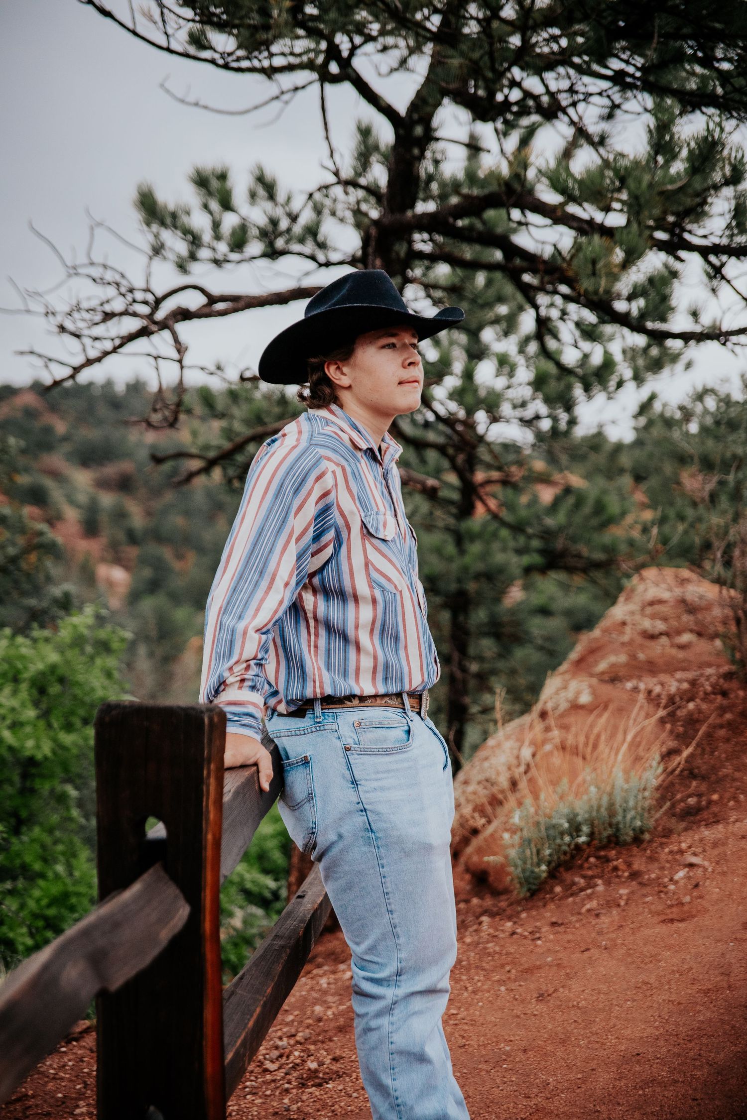 A high school senior in western wear sits contemplatively on red rock steps with wooden railings at Garden of the Gods in Colorado Springs