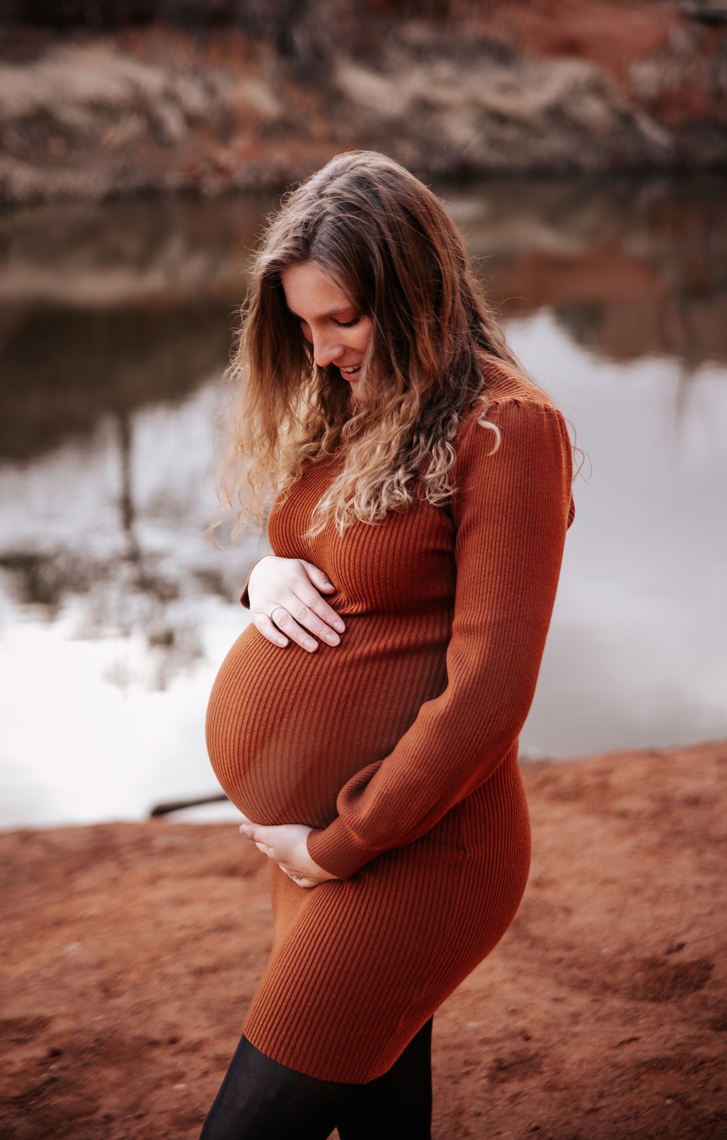 Maternity photos taken against red rocks show tender moments as sun sets over a lake.