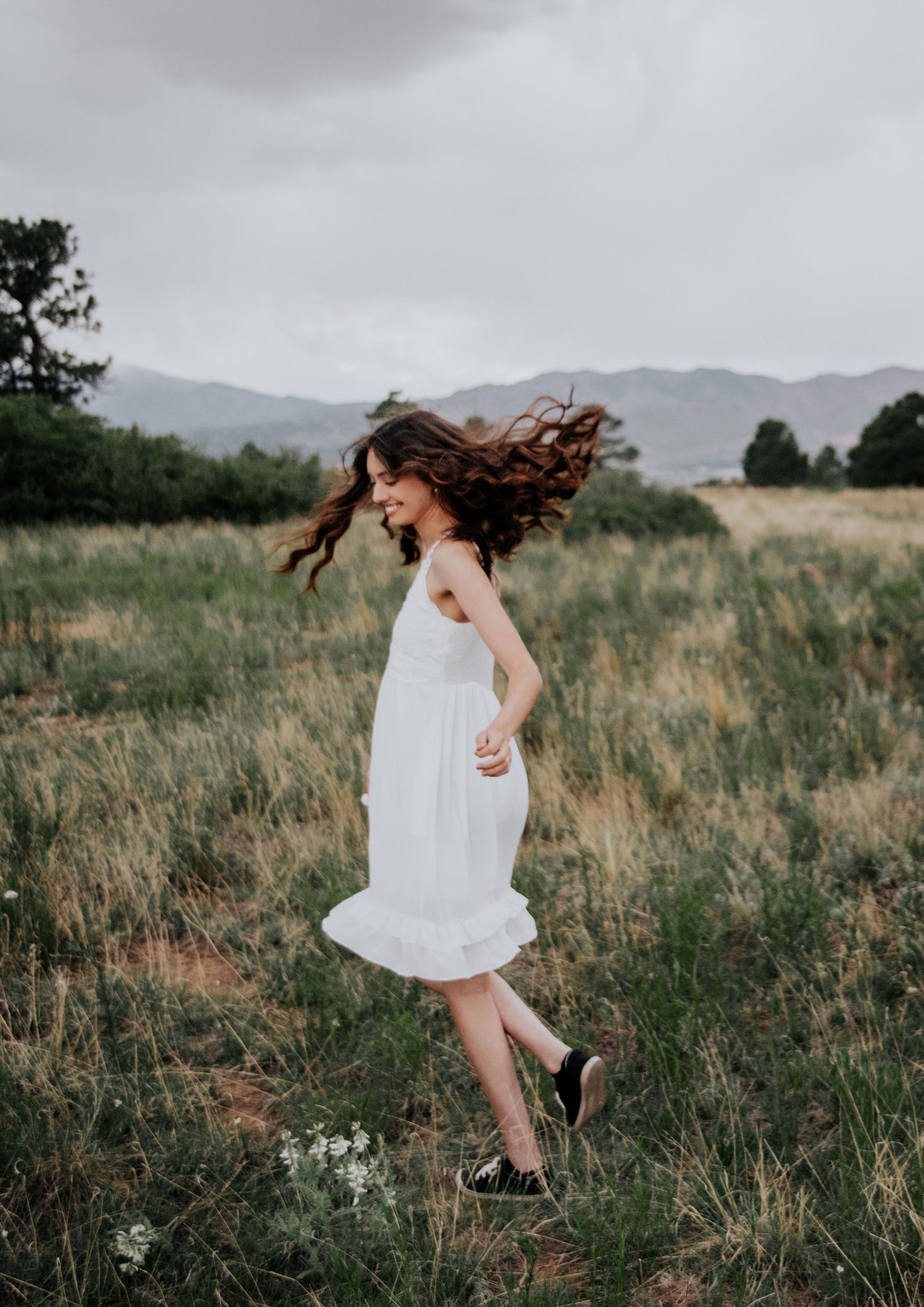 A person in a white dress dances freely in a grassy meadow against a mountainous landscape.