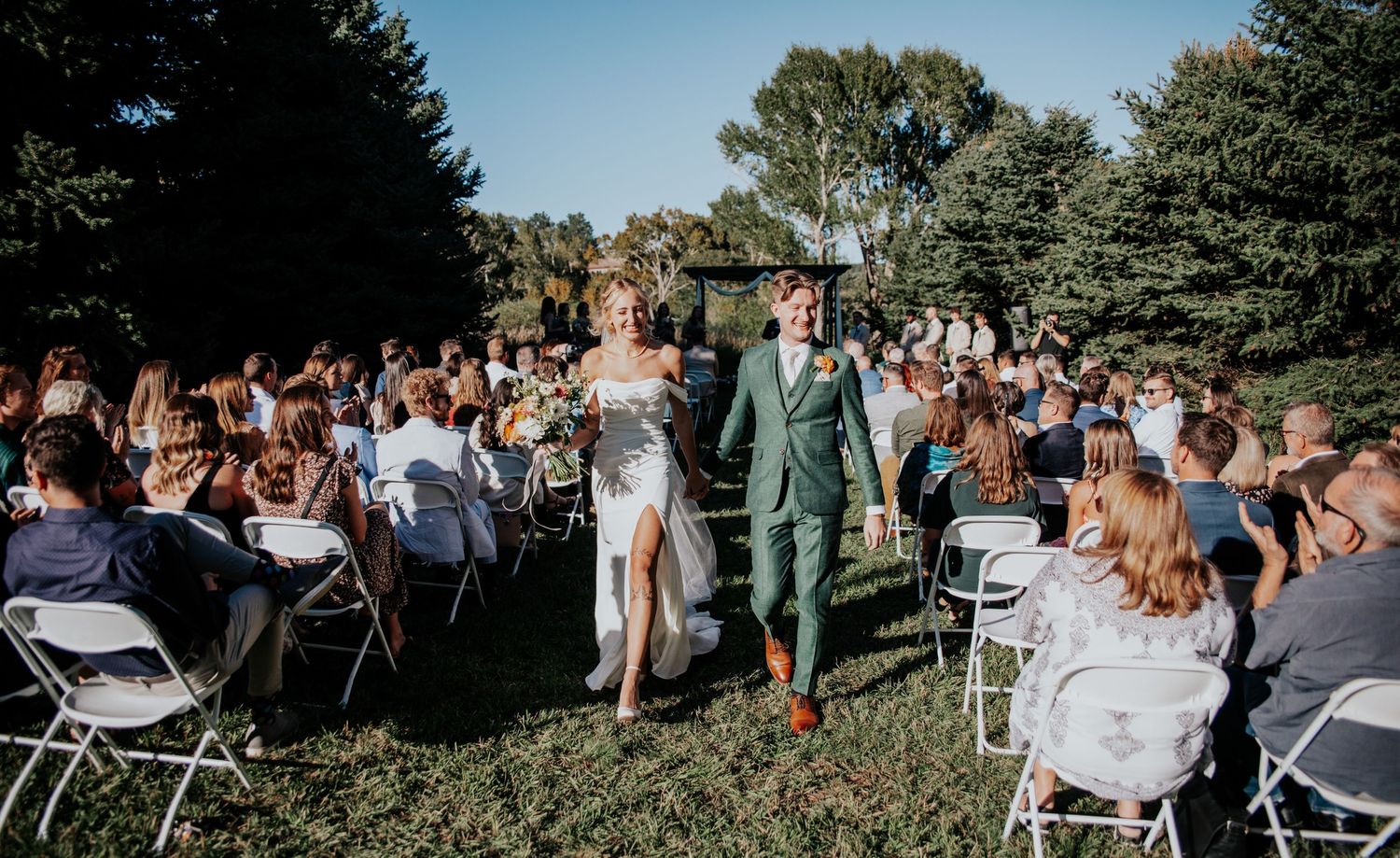 Newlyweds walk down the aisle as guests in white chairs celebrate at an outdoor garden wedding.