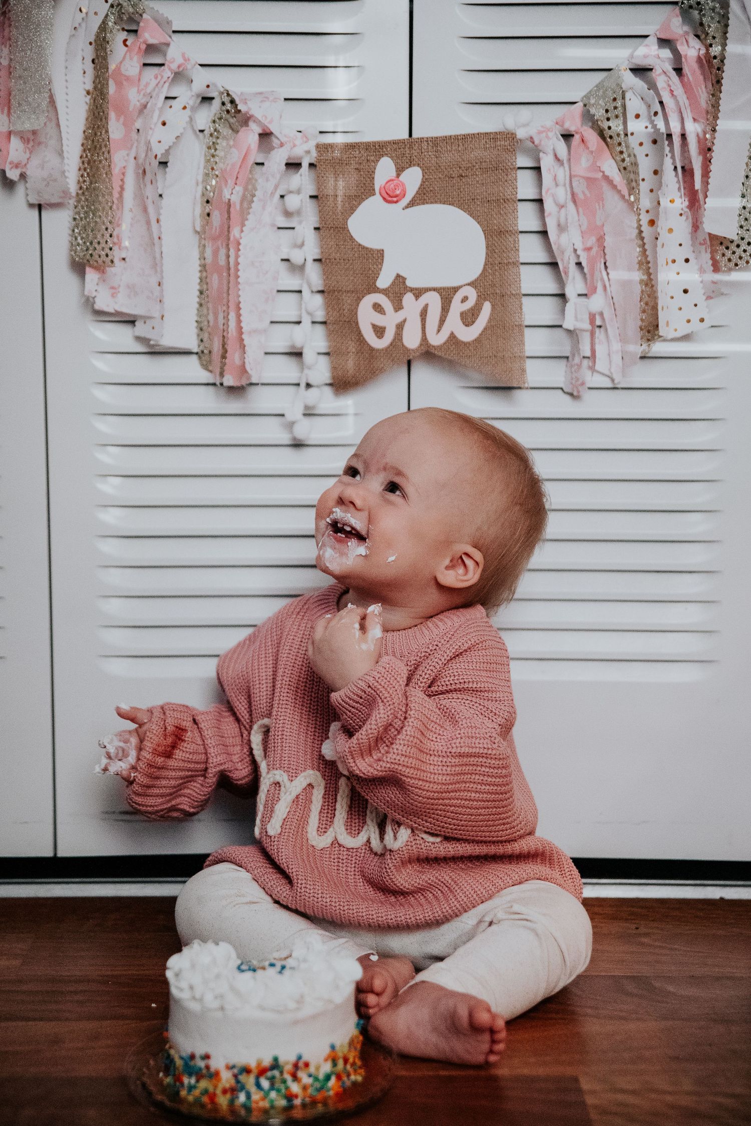 Joyful baby in pink sweater during cake smash photoshoot with white frosted cake.