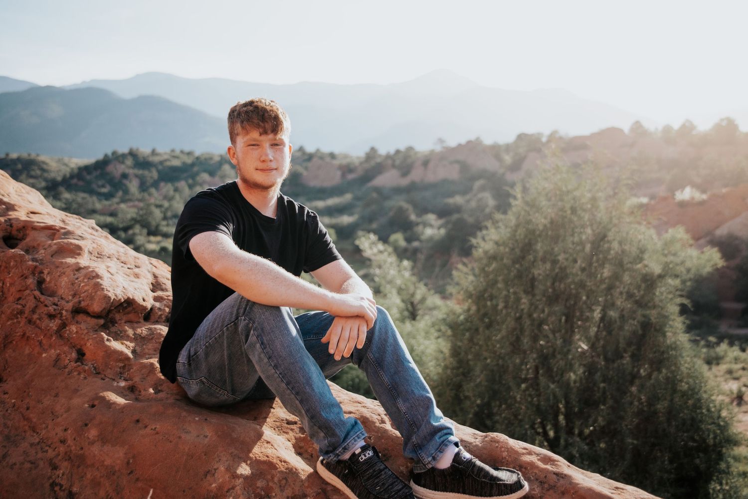 Person in dark clothing sits on red rocks overlooking mountain landscape at sunset.