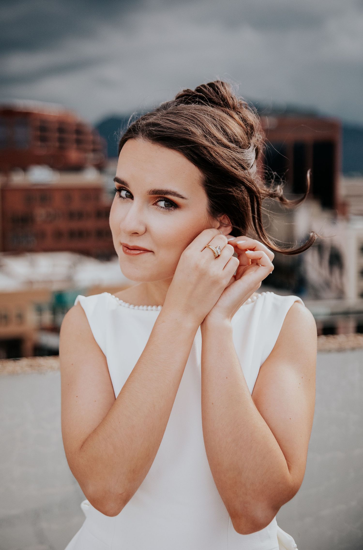Bride in elegant white sleeveless dress adjusting her earring on urban rooftop.